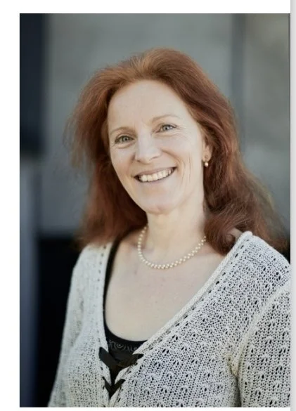 A middle-aged woman with red hair smiling, wearing a white knitted cardigan, a black top, a pearl necklace, standing outdoors with a blurred background.