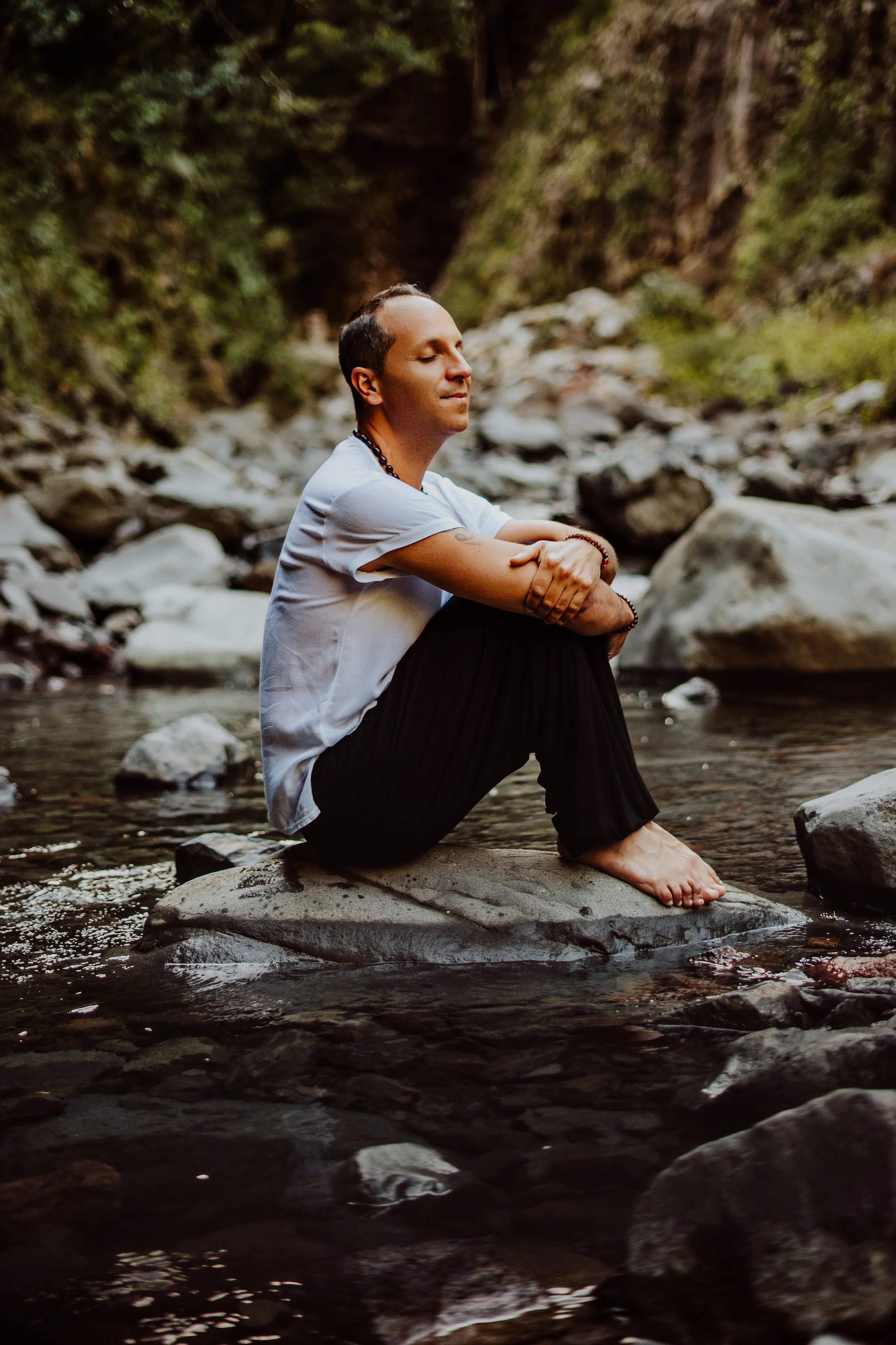 A man sitting cross-legged on a large rock in a creek, with eyes closed in meditation, surrounded by rocks and greenery.