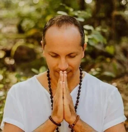 A man with short hair and a white shirt, standing outdoors with a background of greenery, holding his hands together in a prayer position and looking down with a peaceful expression.