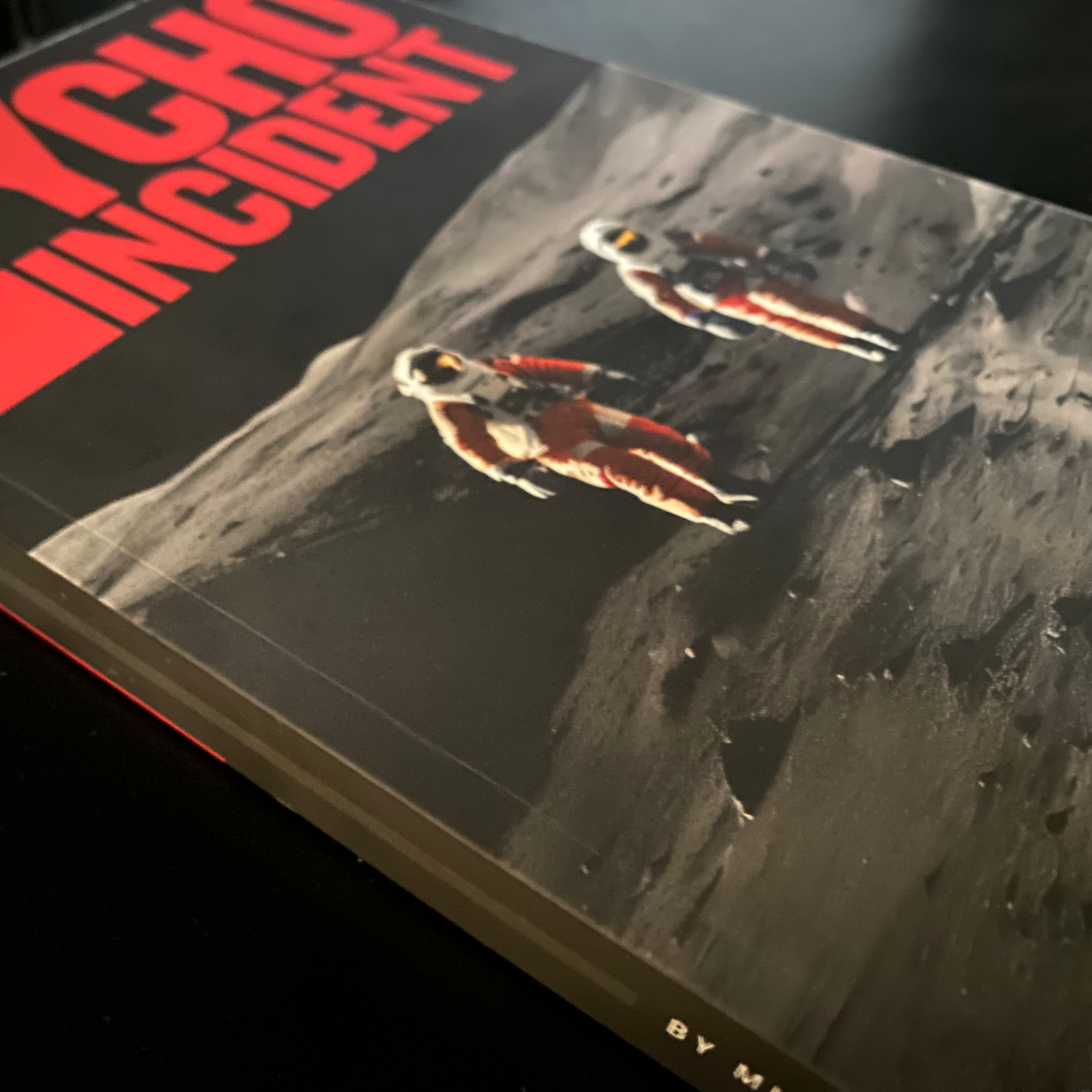 A book titled "The Event” with a photograph of two astronauts walking on the moon’s surface, wearing orange suits, with lunar landscape in the background.
