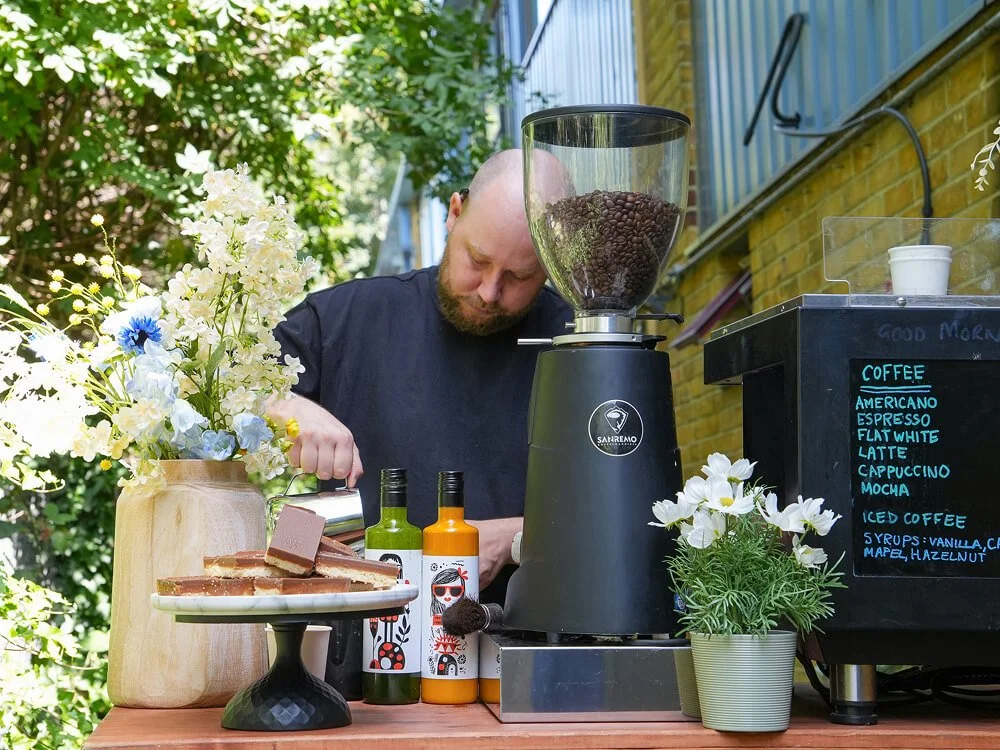 Joe making coffee behind grace&skye's mobile coffee bar on a sunny day. There are flowers arranged on the bar as well as shortbread and syrups