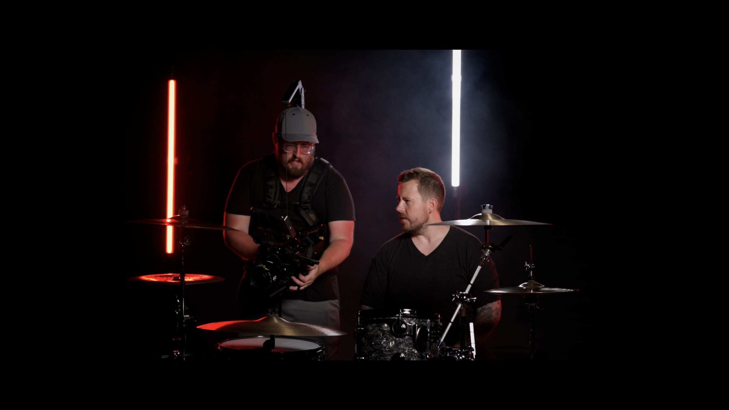 Two men with drums in a dark studio illuminated by vertical red and white neon lights, one man is seated at the drums while the other stands next to him holding a camera.