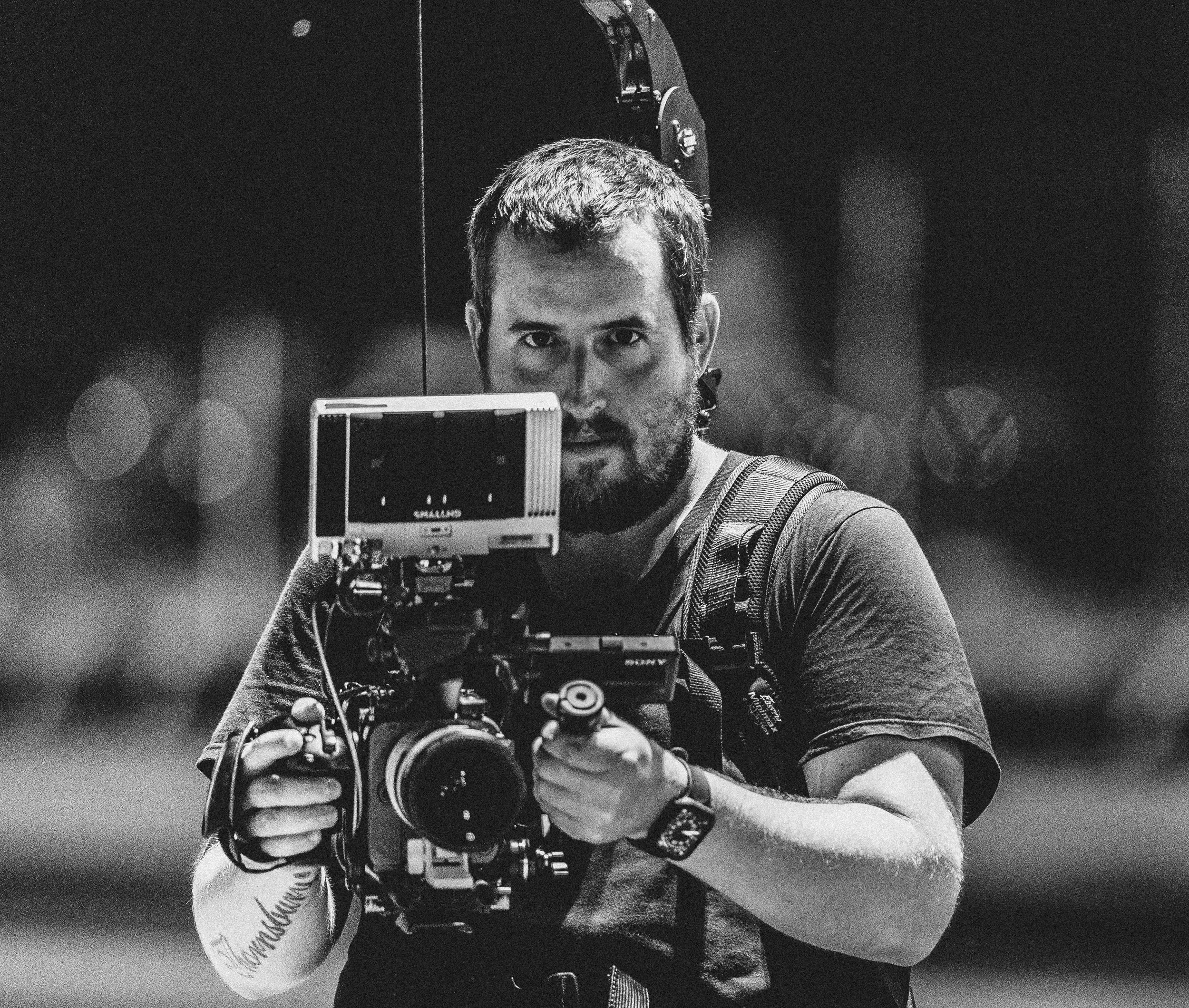 A man with short hair and a beard holding a professional video camera, looking directly at the camera in a black and white photograph.