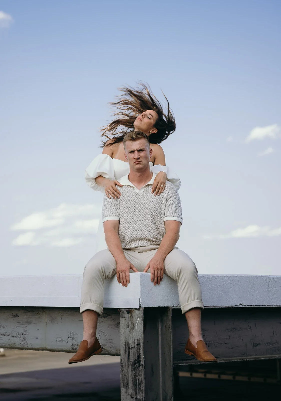 Couple in monochromatic cream. The man is sitting on the edge of a building as his fiance whips her hair behind him.