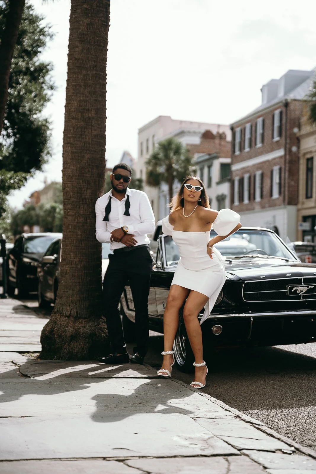 Woman in white dress leaning against a vintage mustang. Man leaning against a palm tree on Broad Street in Charleston, SC.