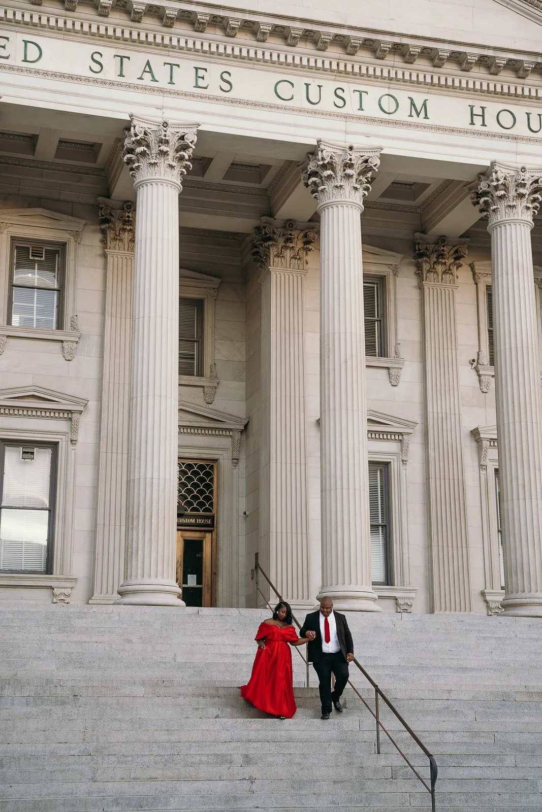 Fiances walking down the steps. The man is wearing a black suit with a red tie. The woman is wearing a long formal red dress at the United States Custom House in Charleston, SC.