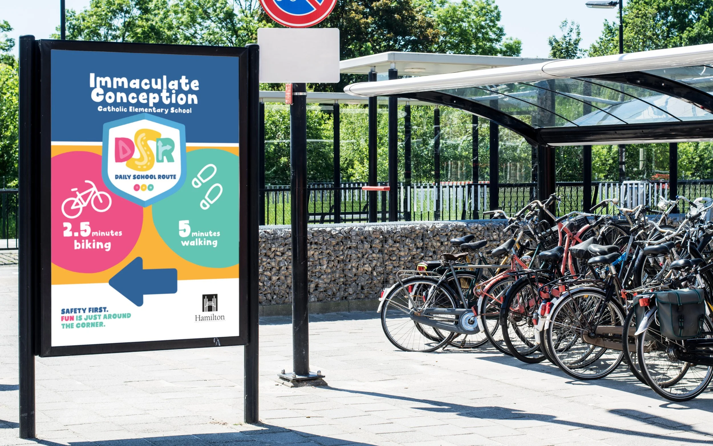 Outdoor bicycle parking area with a sign promoting "Immaculate Conception Catholic Elementary School" featuring daily school routes, 2.5 minutes biking and 5 minutes walking options, with an emphasis on safety.