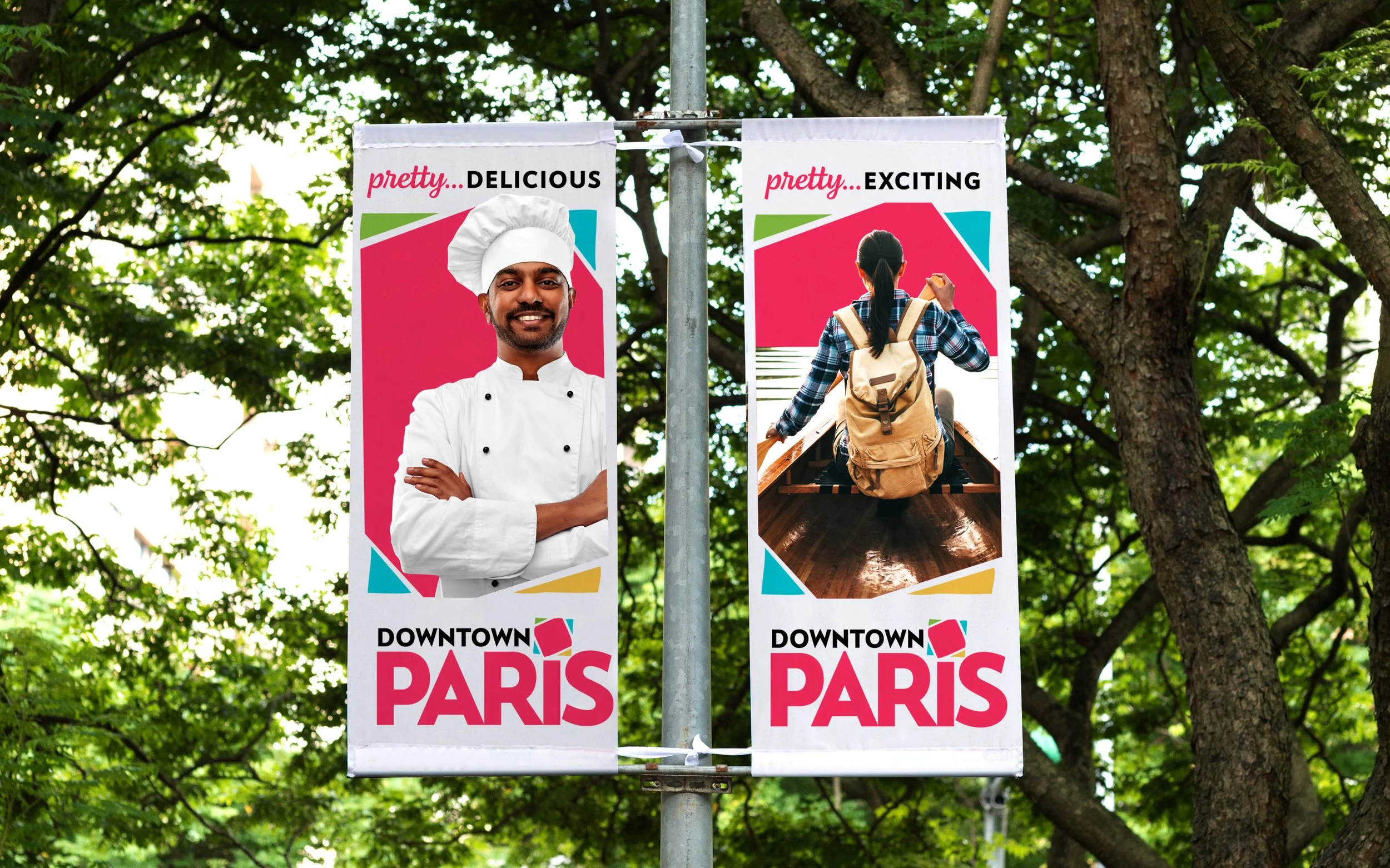 Outdoor banners promoting Downtown Paris, featuring a smiling chef on the left and a person with a backpack on the right, surrounded by trees.