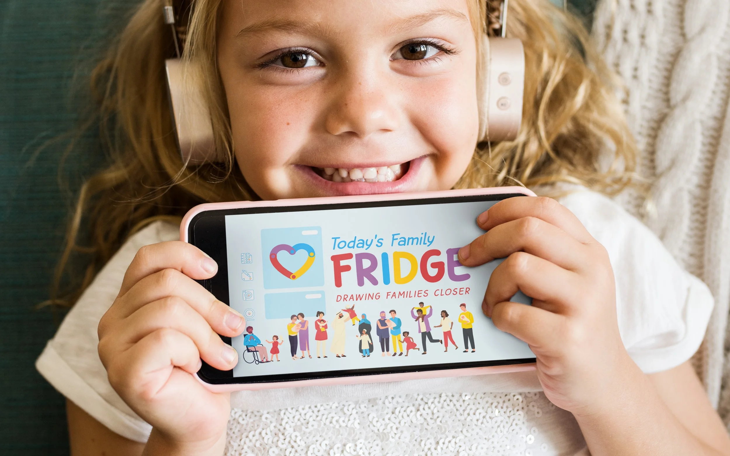 A child wearing headphones smiles while holding a phone displaying an app called "Today's Family Fridge: Drawing Families Closer." The app features colorful illustrations of diverse families.