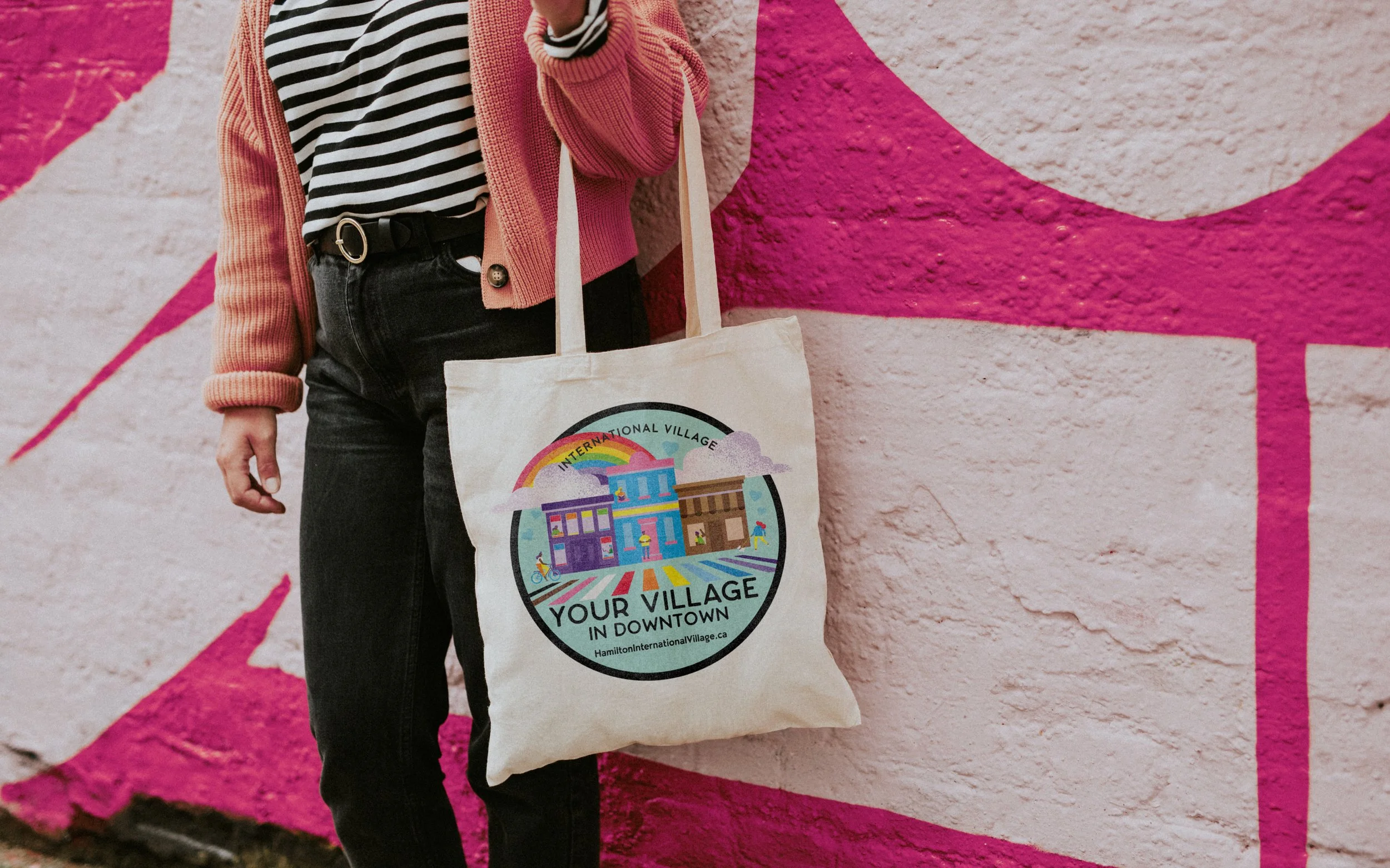 Person holding a tote bag with "International Village" design against a pink mural background.