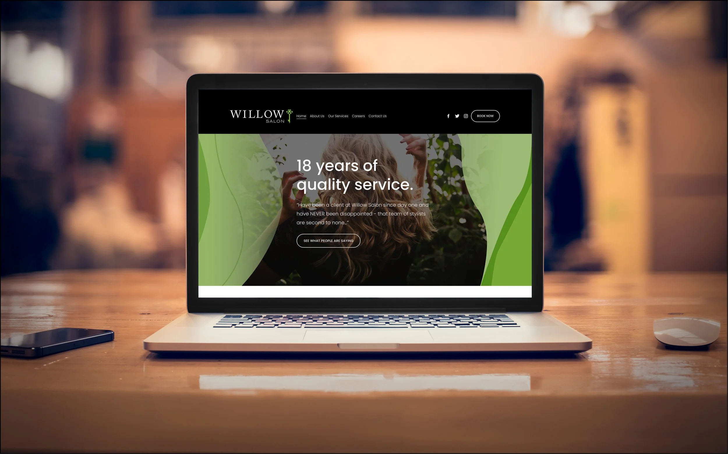 Laptop screen displaying the Willow Salon website with text about 18 years of quality service, placed on a wooden desk with a smartphone and mouse nearby.