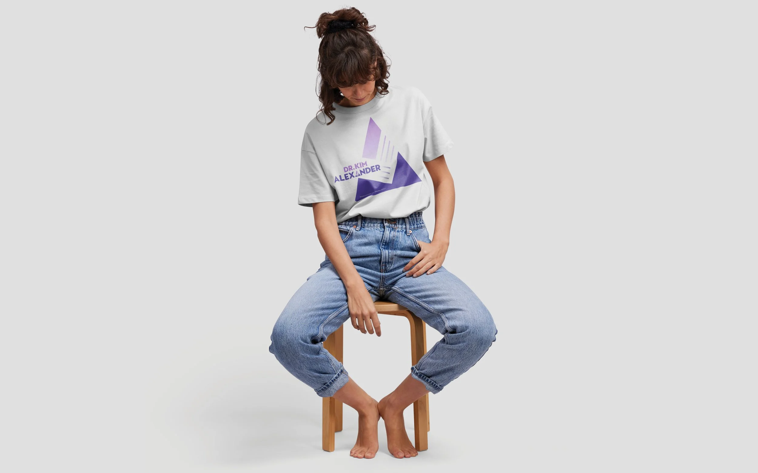 Person sitting on a stool wearing a white graphic T-shirt with "DR. KIM ALEXANDER" and blue jeans, barefoot, looking down.