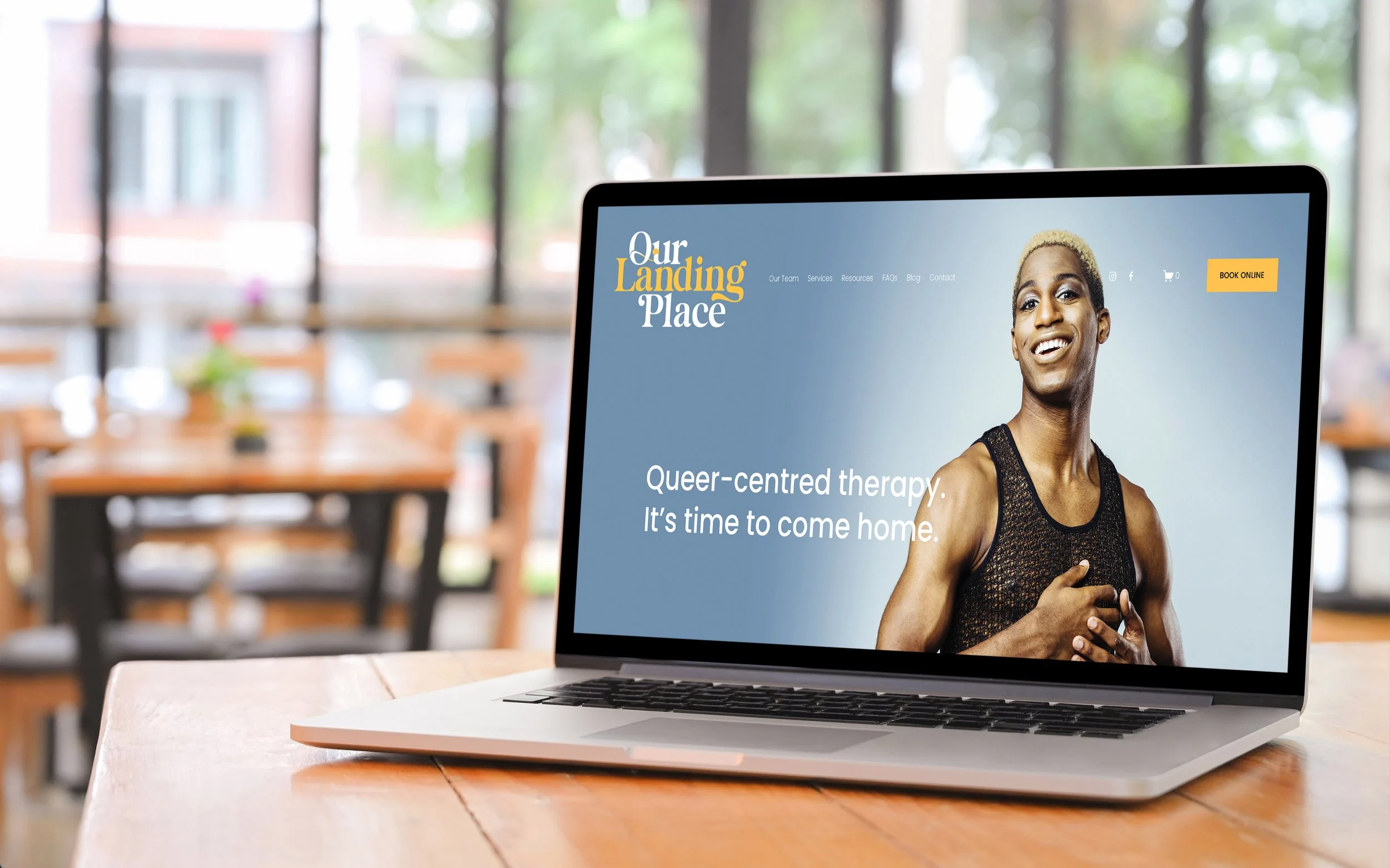 Laptop displaying a website for "Our Landing Place" promoting queer-centered therapy, with an image of a smiling person in a tank top. The text reads, "Queer-centred therapy. It’s time to come home." The background shows a blurred cafe setting.