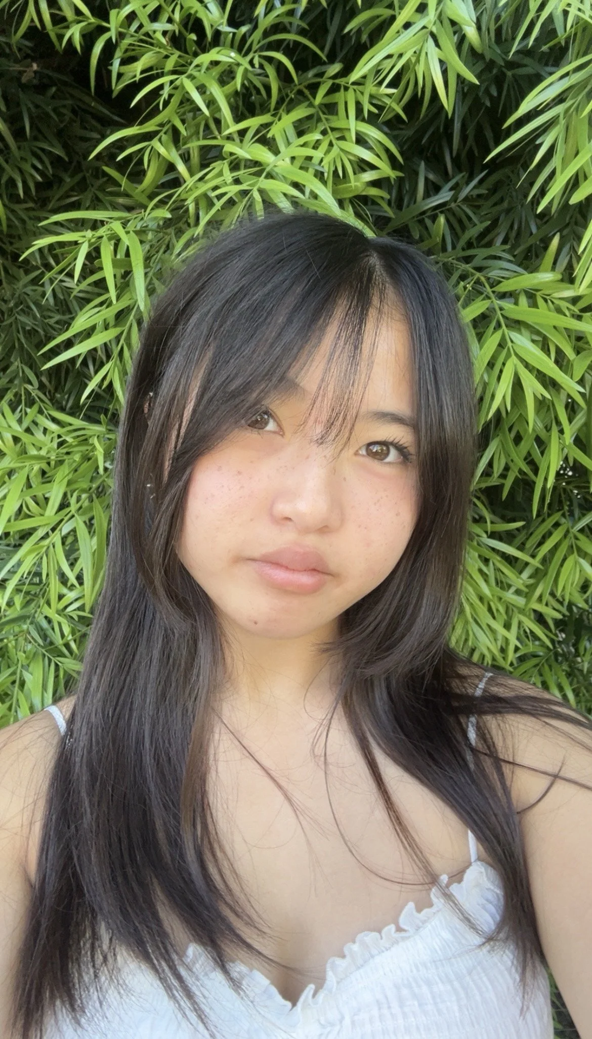 A young woman with dark hair and light skin, standing in front of green foliage, wearing a white top with thin straps.