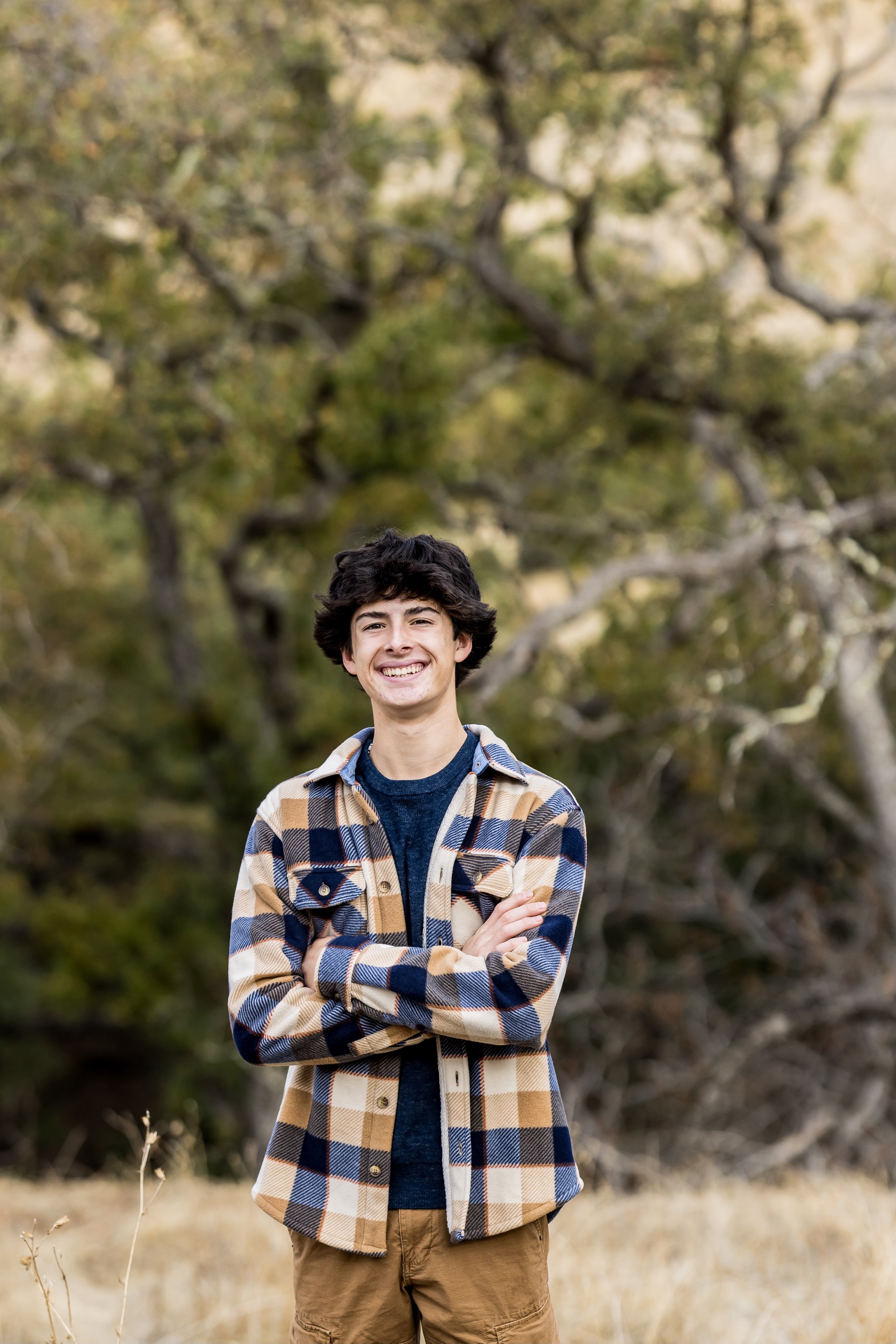 A young man with dark, curly hair standing outdoors in front of a background of trees with green and yellow leaves, smiling and crossing his arms, wearing a plaid jacket over a dark shirt and tan pants.