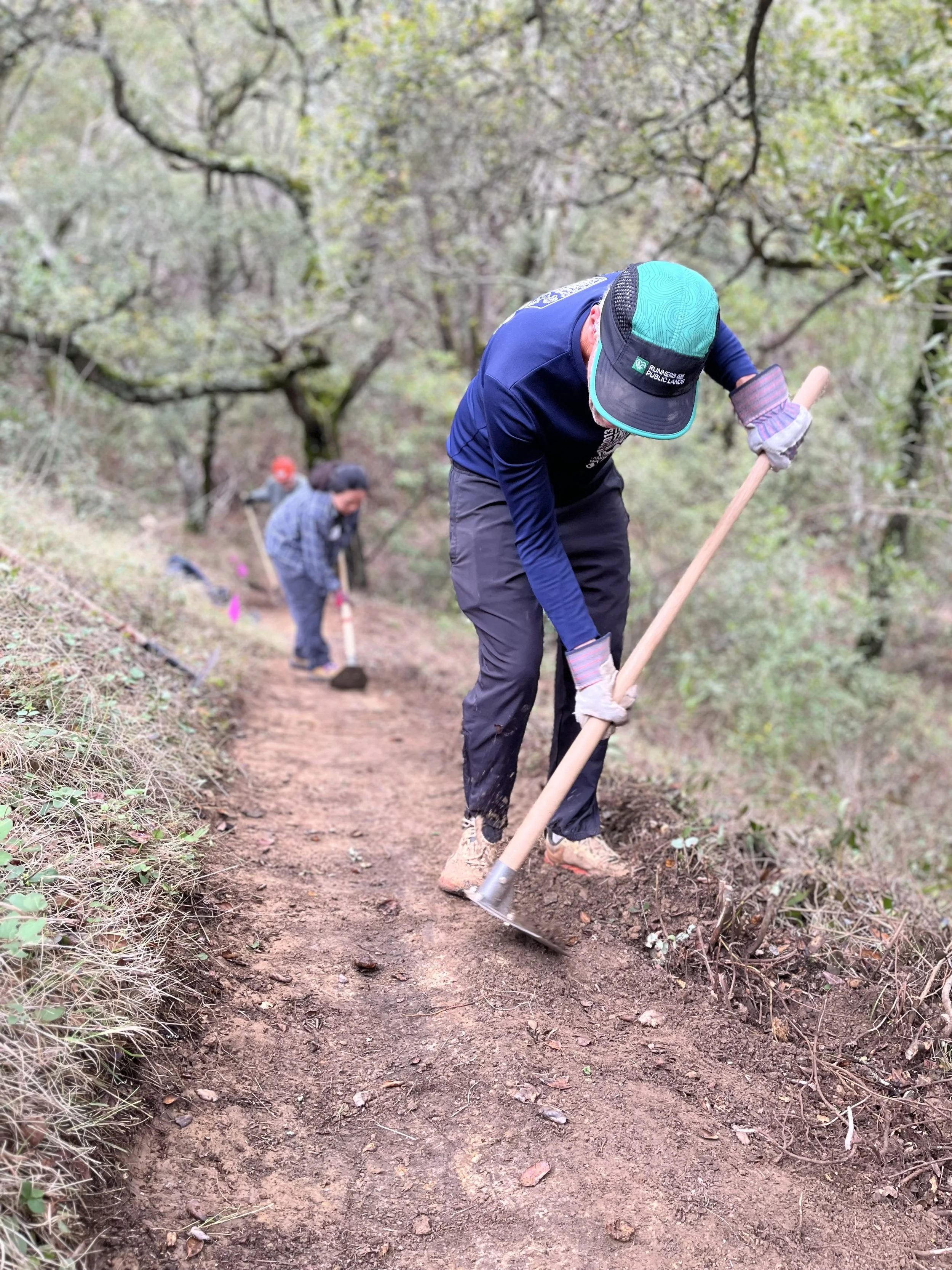 Two people working together to clear a dirt trail in a forest, one with a shovel in the foreground and the other with a tool in the background.
