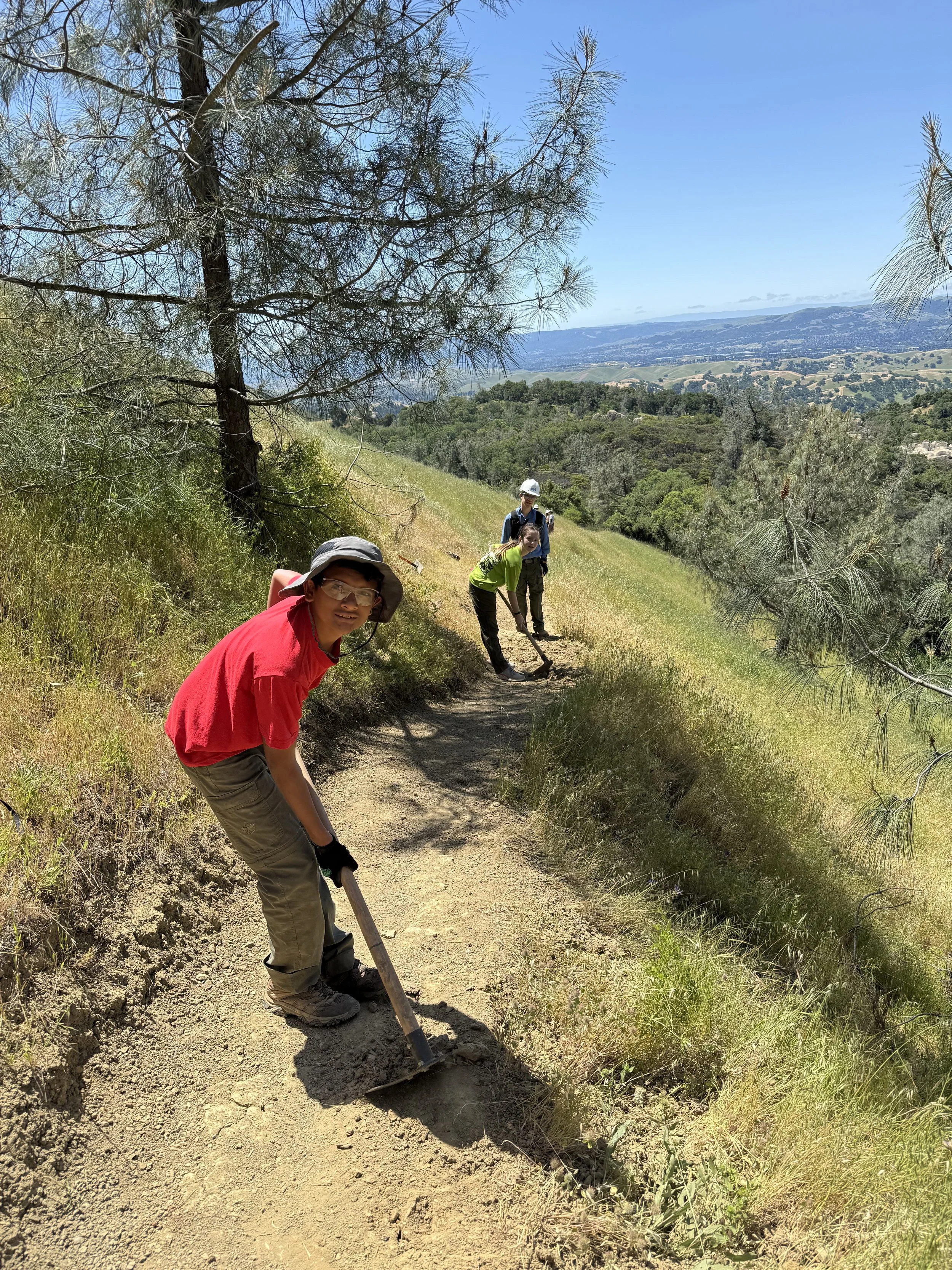 Three people working on trail maintenance on a dirt path in a hilly, wooded area under a blue sky