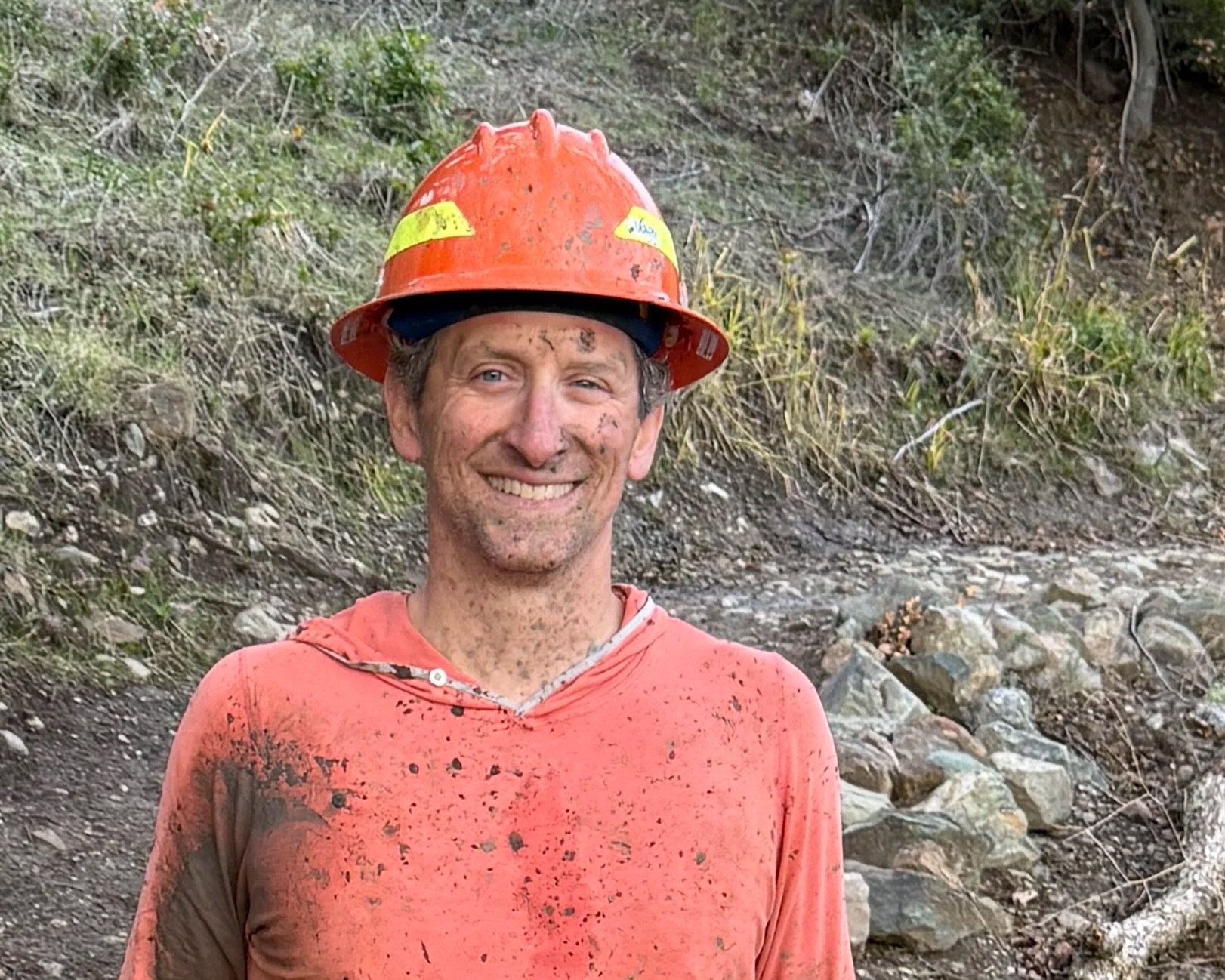 A smiling man wearing a plaid shirt and a cap outdoors with a black puppy, in front of a scenic autumn landscape of colorful trees and mountains.