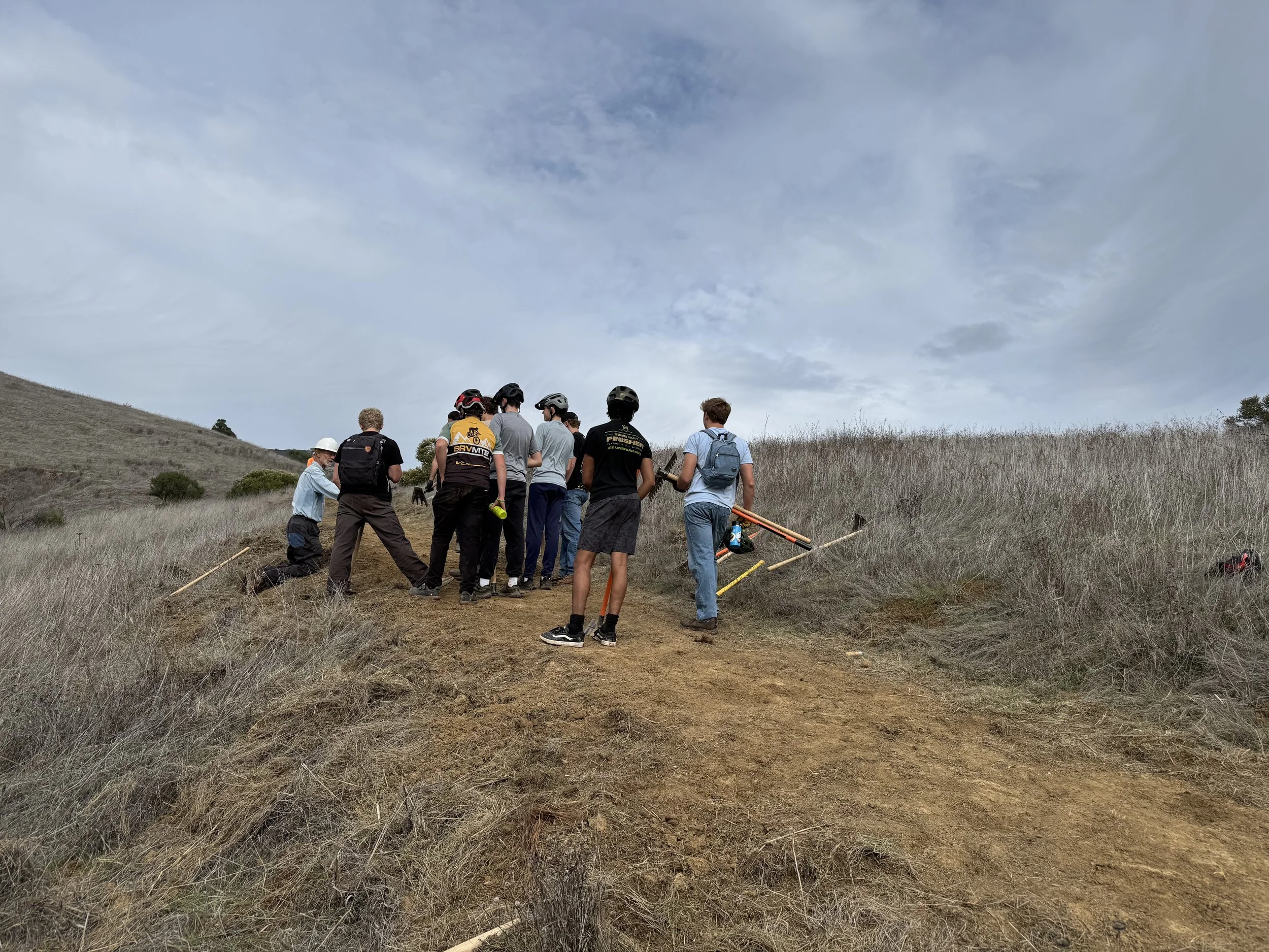 A group of people, some wearing helmets and backpacks, gathered on a dirt trail in a hilly, grassy area, possibly preparing for a hike or outdoor activity, under a partly cloudy sky.