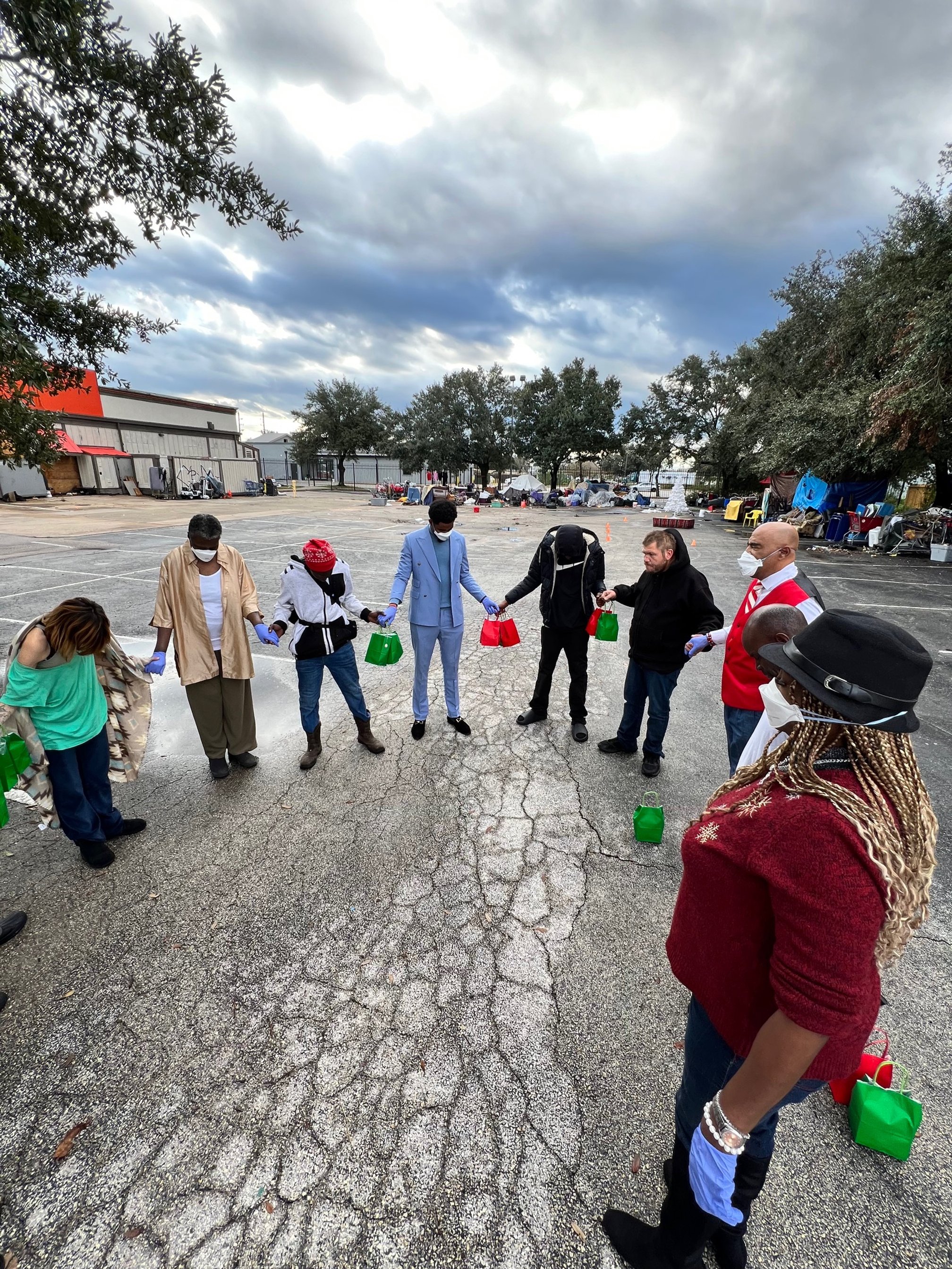 A group of people standing in a circle outdoors, holding hands with a parking lot and trees in the background, and some tents and items scattered around.
