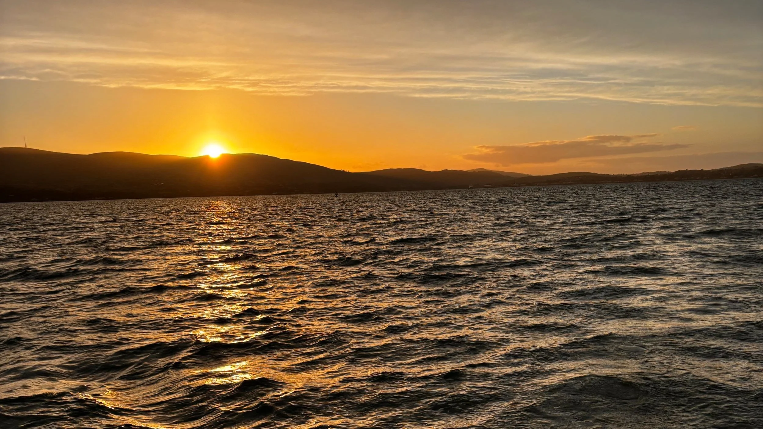 Sunset over a large body of water with gentle waves, silhouette of hills in the background, and a colorful sky with clouds.