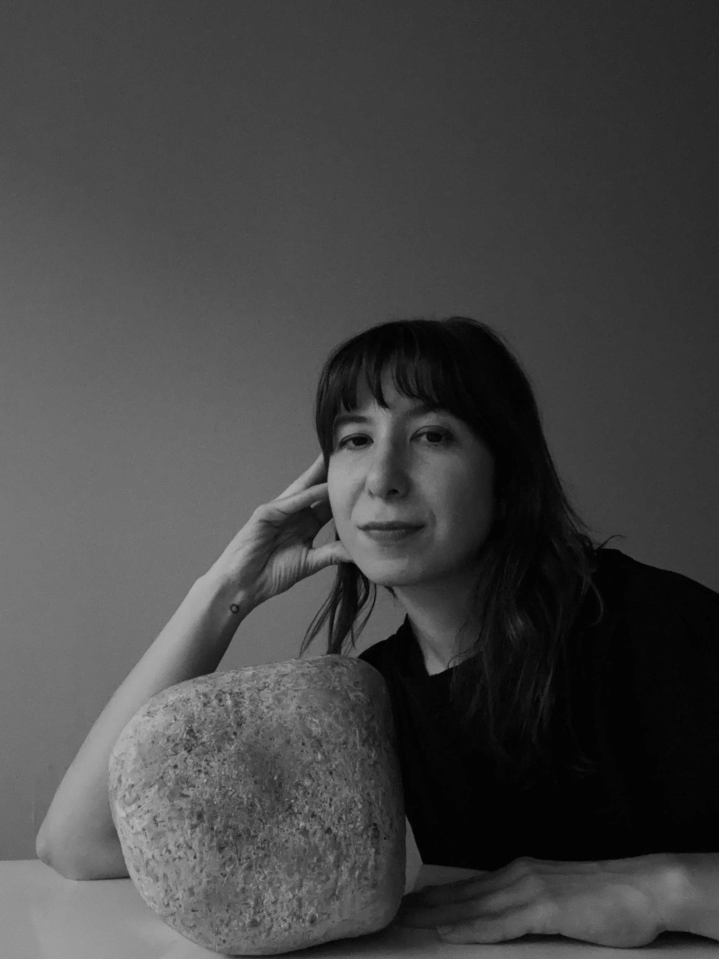 Black and white portrait of a woman sitting at a table, resting her head on her hand, next to a rounded rock or stone, with a plain background.