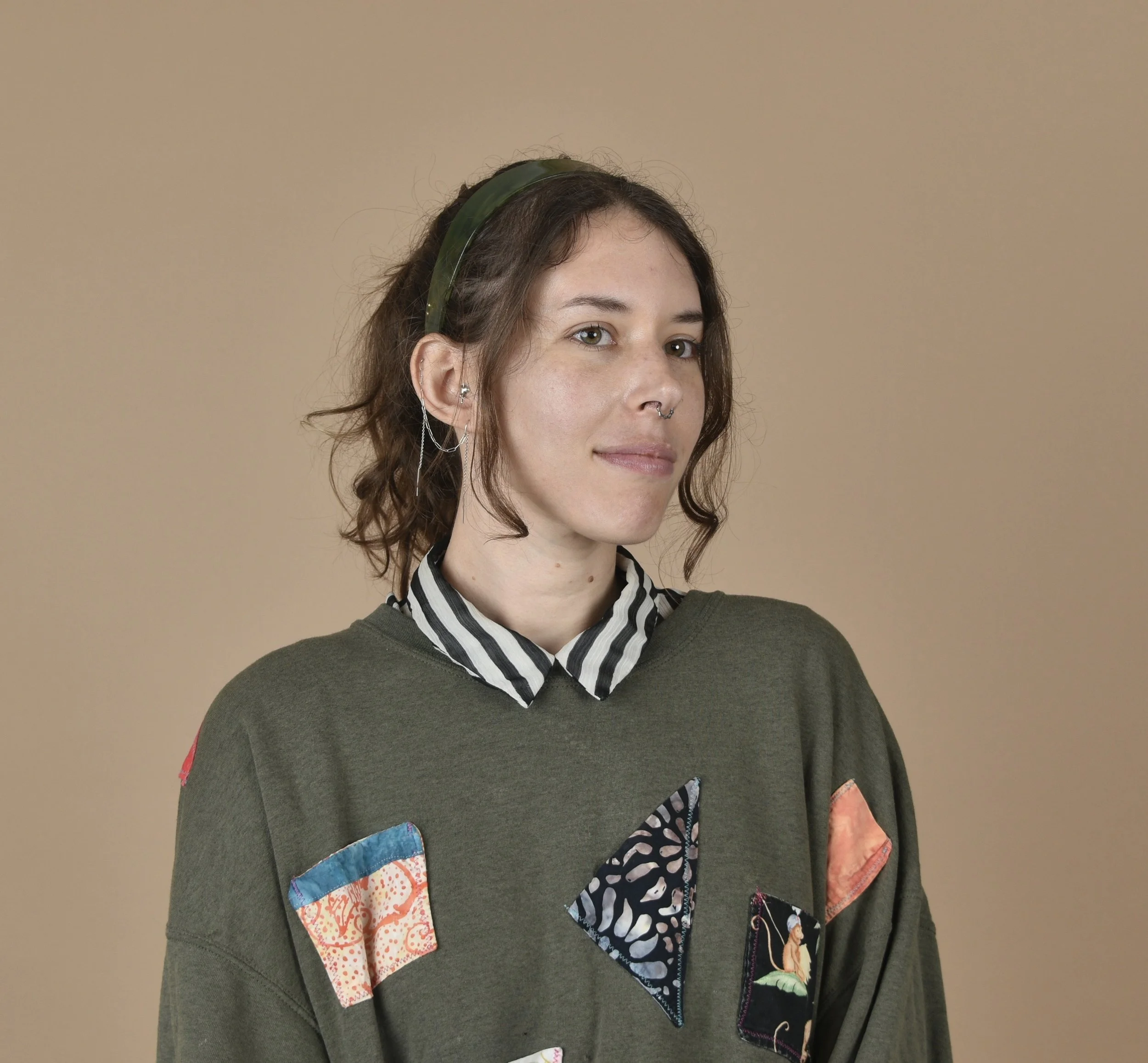 A young woman with shoulder-length curly brown hair wearing a green sweatshirt with patchwork designs and a striped collared shirt underneath. She has multiple earrings and a nose ring, and a green headband in her hair, standing against a beige background.