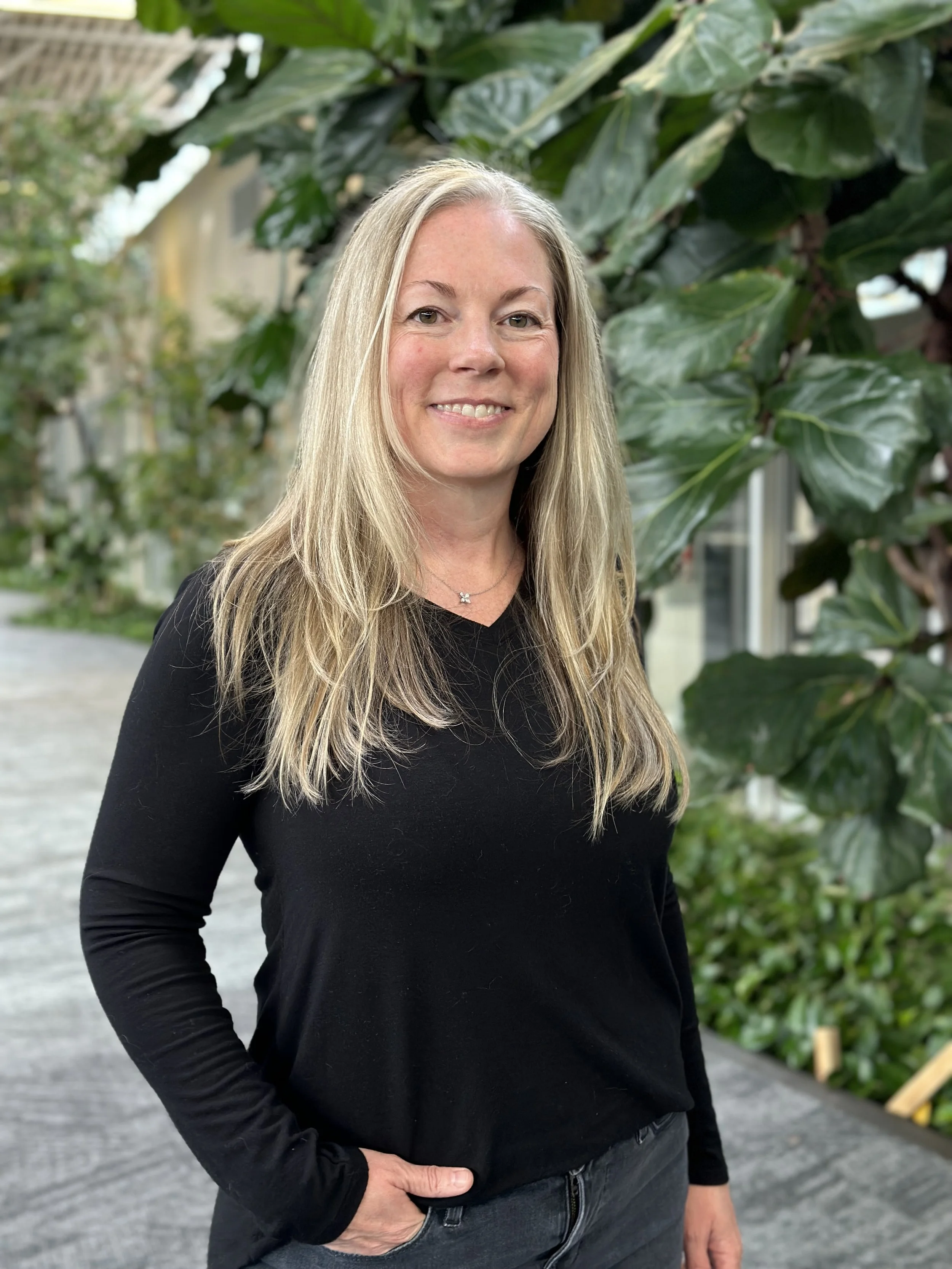 A woman with long blonde hair, smiling and crossing her arms, standing in front of large green leaves of a plant, outside or in a garden setting.