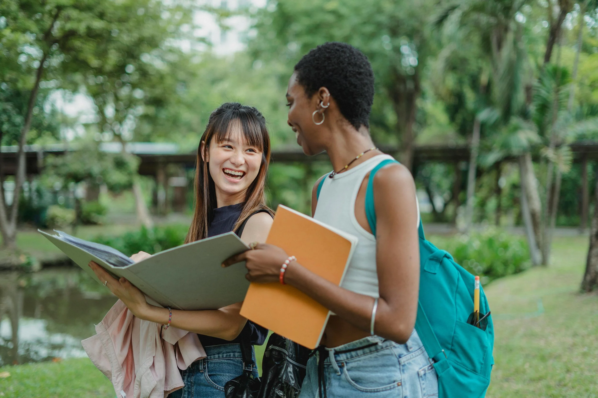 Two young women smiling and talking outdoors in a park with trees, holding notebooks and folders.