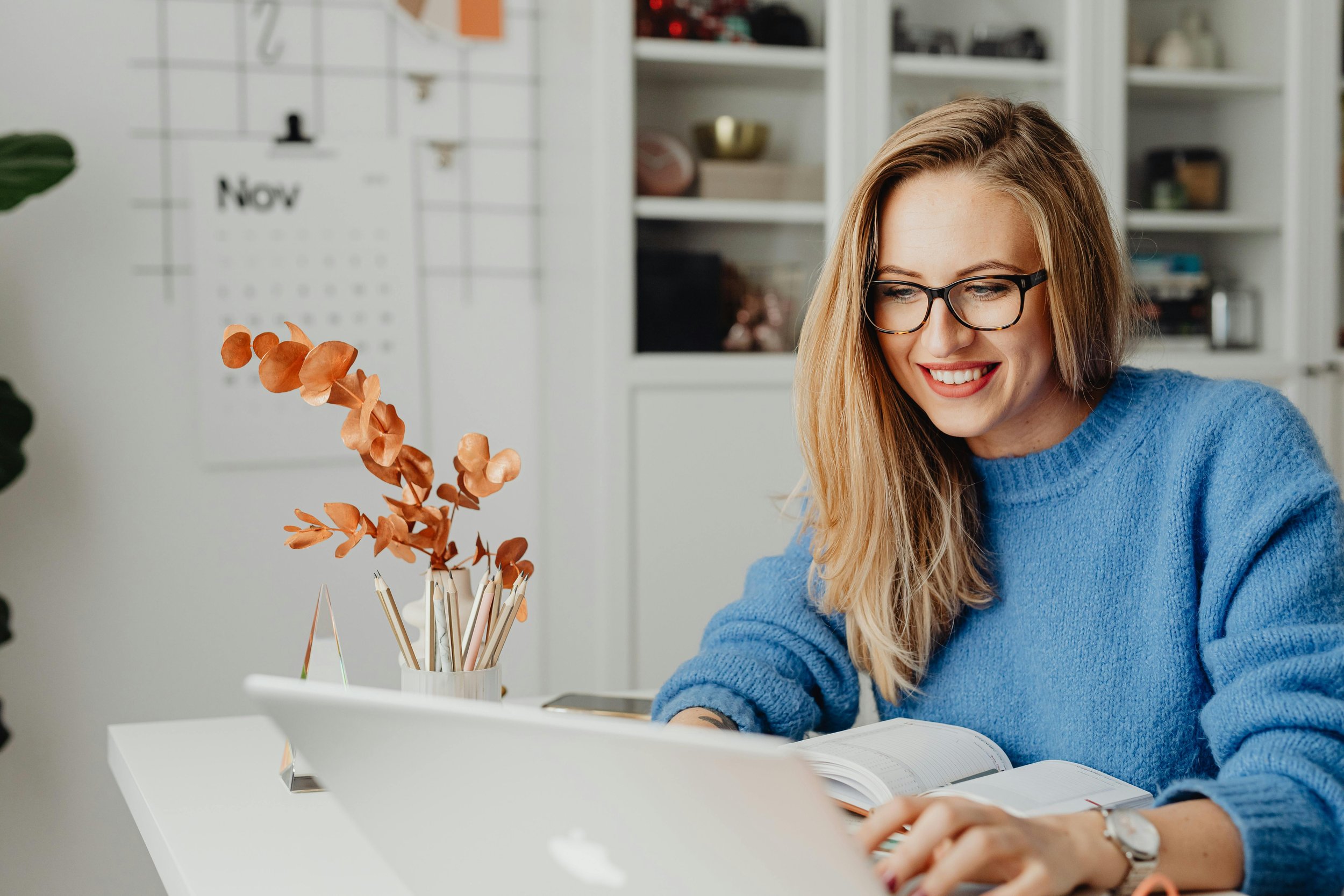 A woman with blonde hair and glasses wearing a blue sweater working on a laptop at a white desk with a notebook and a cup filled with dried orange leaves and pens in it, in a bright room with white shelves and a calendar in the background.