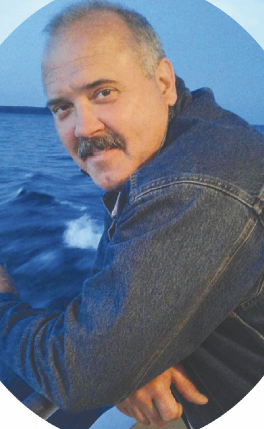 A man with a mustache and short hair leaning on a boat railing, with the ocean and a blue evening sky in the background.