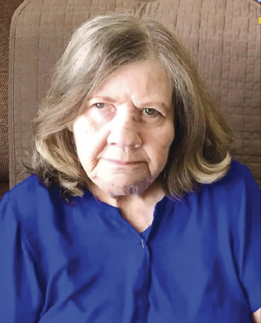 A woman with shoulder-length gray hair wearing a blue shirt, sitting indoors with a wooden background.