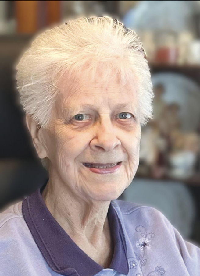 An elderly woman with white hair and blue eyes smiling, wearing a purple shirt, with a blurred background of a room with shelves and decorative items.