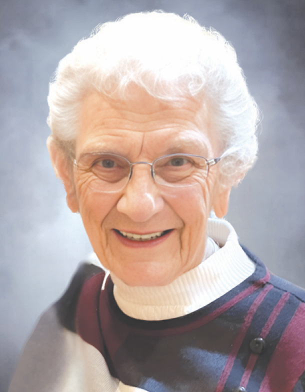 Close-up portrait of an elderly woman with white curly hair, wearing glasses, a white turtleneck, and a color-blocked sweater, smiling at the camera against a neutral background.