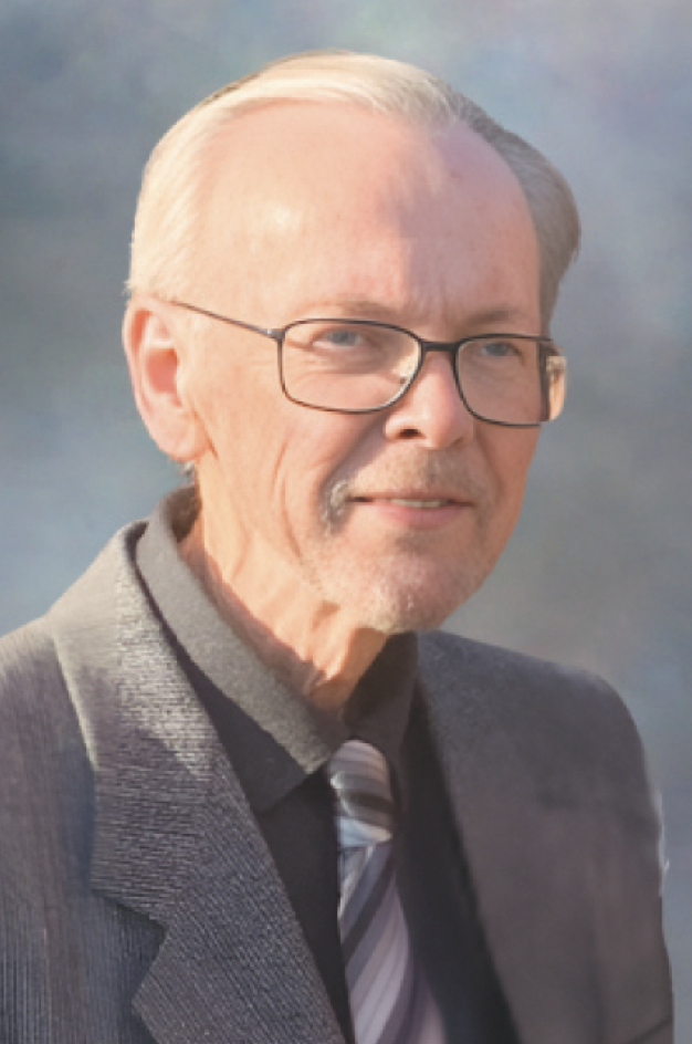 A man with white hair, glasses, and a beard, wearing a gray suit and a striped tie, standing outdoors with a mountainous background.