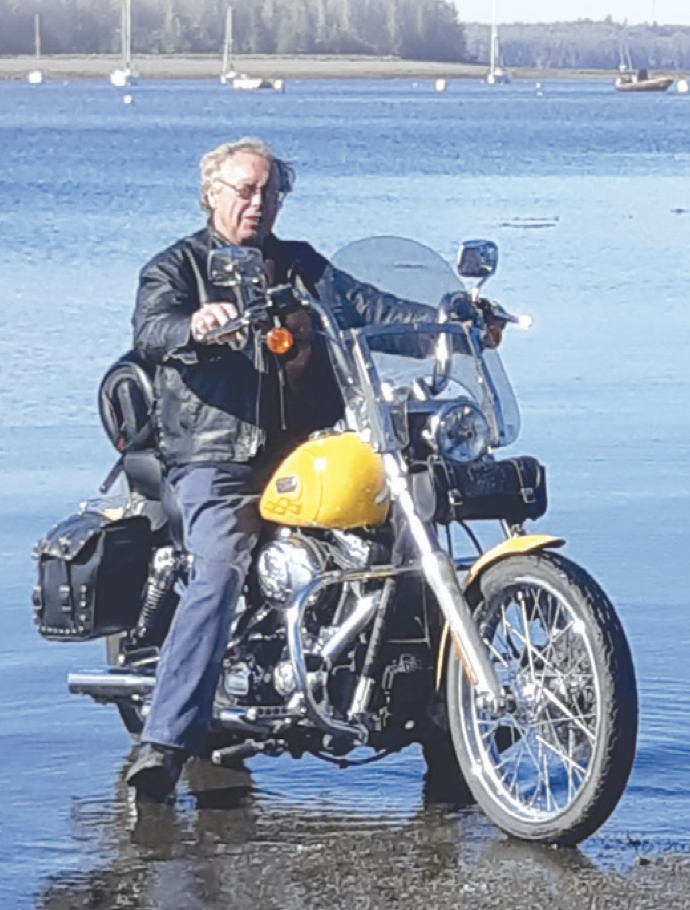 A man in a black leather jacket and jeans riding a yellow motorcycle in shallow water near a lake with boats and trees in the background.