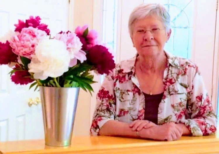 An elderly woman sitting at a dining table with a large bouquet of pink and white peonies in a silver vase.