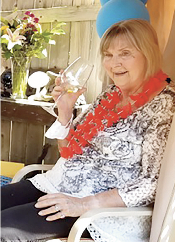 An elderly woman celebrating a birthday with a blue party hat, red lei, and holding a glass of wine, seated outdoors with a bouquet of flowers and a wooden fence in the background.