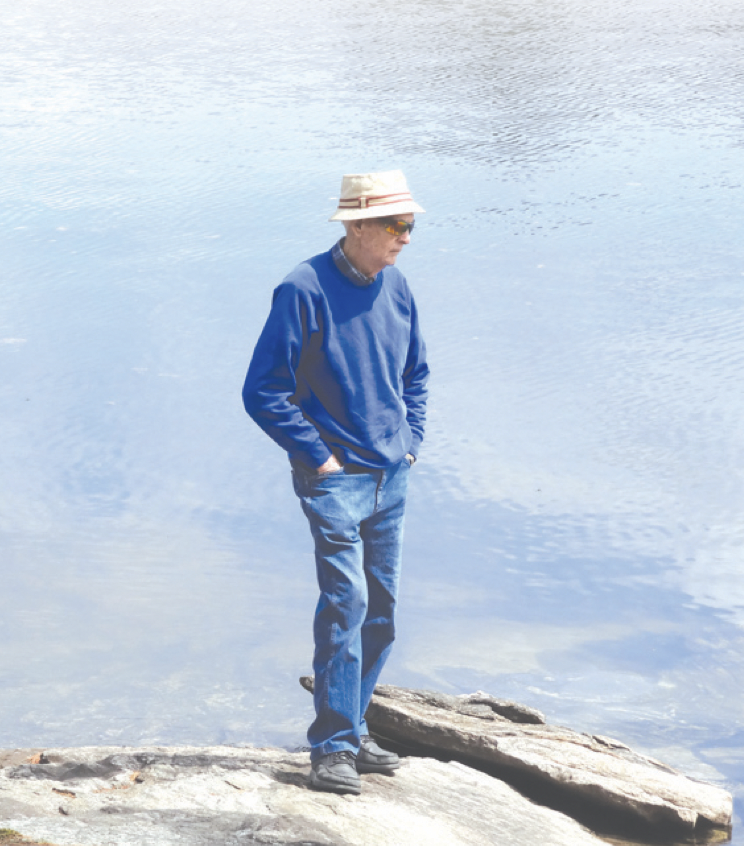 A man wearing a blue sweatshirt, jeans, and a bucket hat standing on a rock by a body of water, looking down with his hands in his pockets.