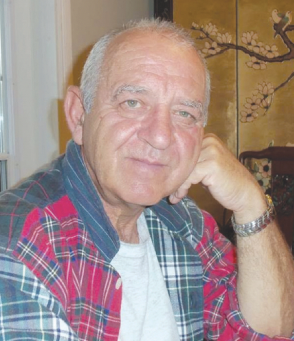 Close-up of an elderly man with short gray hair, wearing a plaid shirt and a watch, resting his face on his hand, in a room with a window and Asian-style art on the wall.