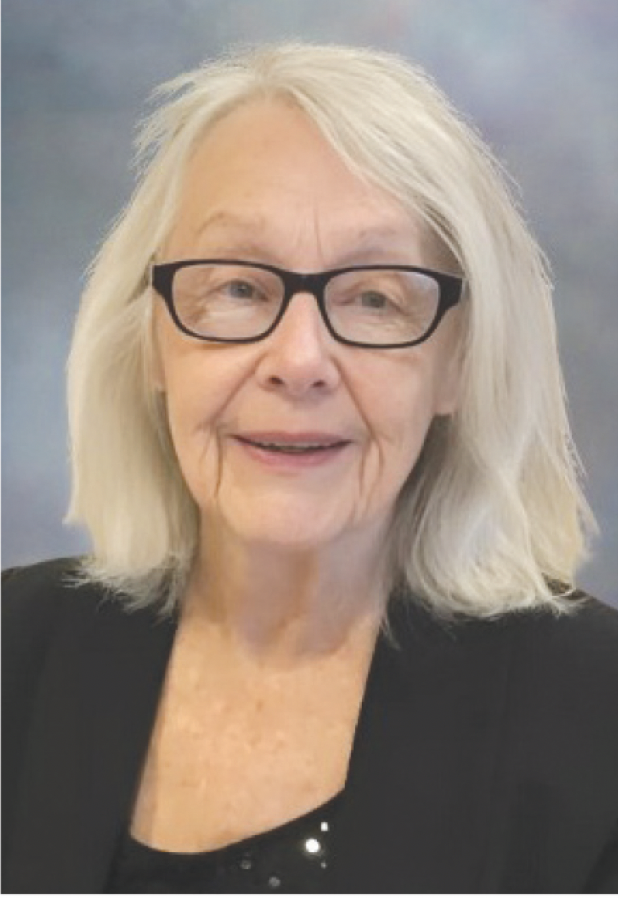 An elderly woman with shoulder-length white hair, wearing black glasses and a black blazer, smiling at the camera against a blurred multi-colored background.