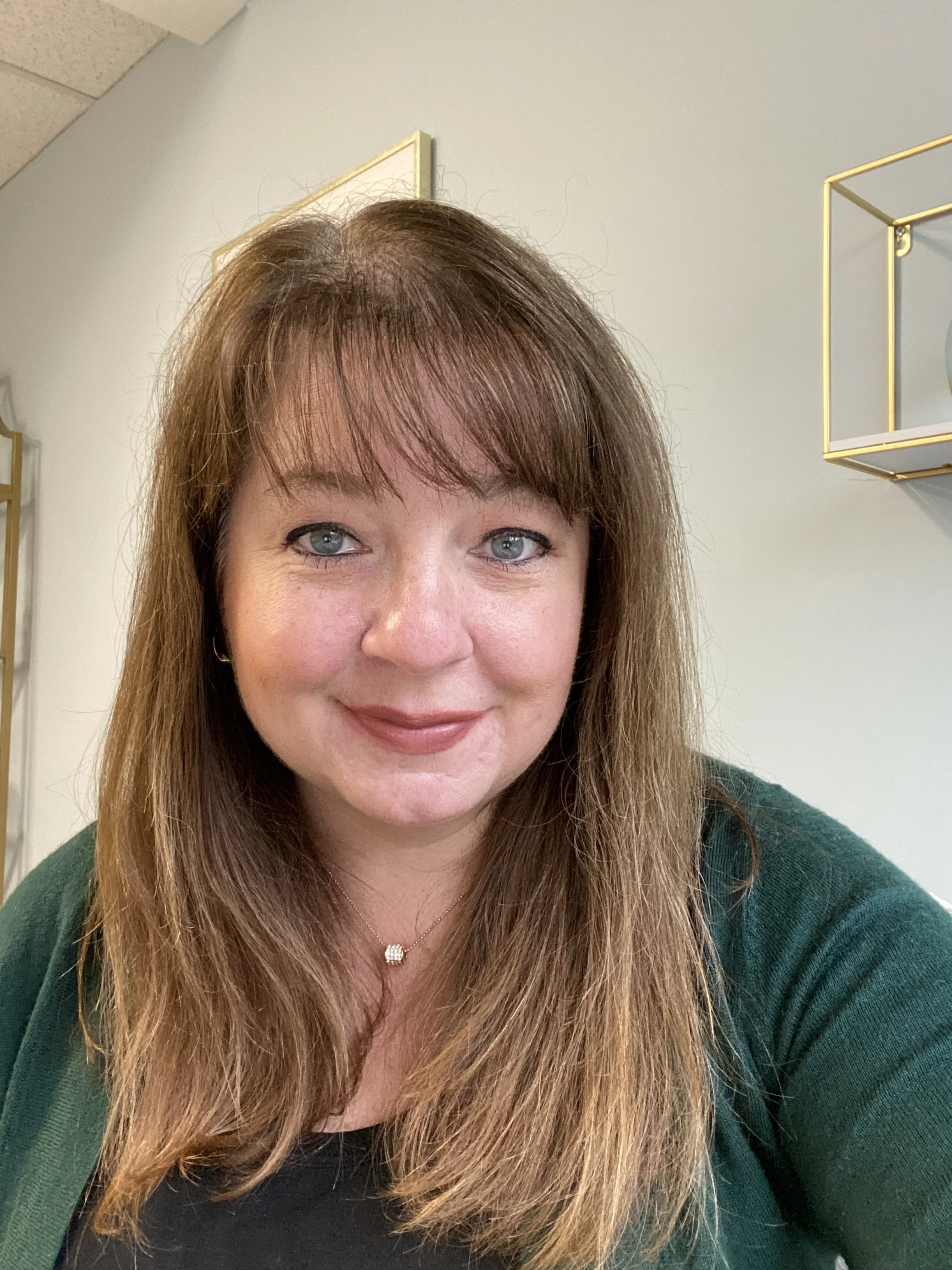 Close-up of a smiling woman with long brown hair and blue eyes, wearing a black top, a green sweater, and a pearl necklace, in an indoor setting with gray walls and gold-framed decor.