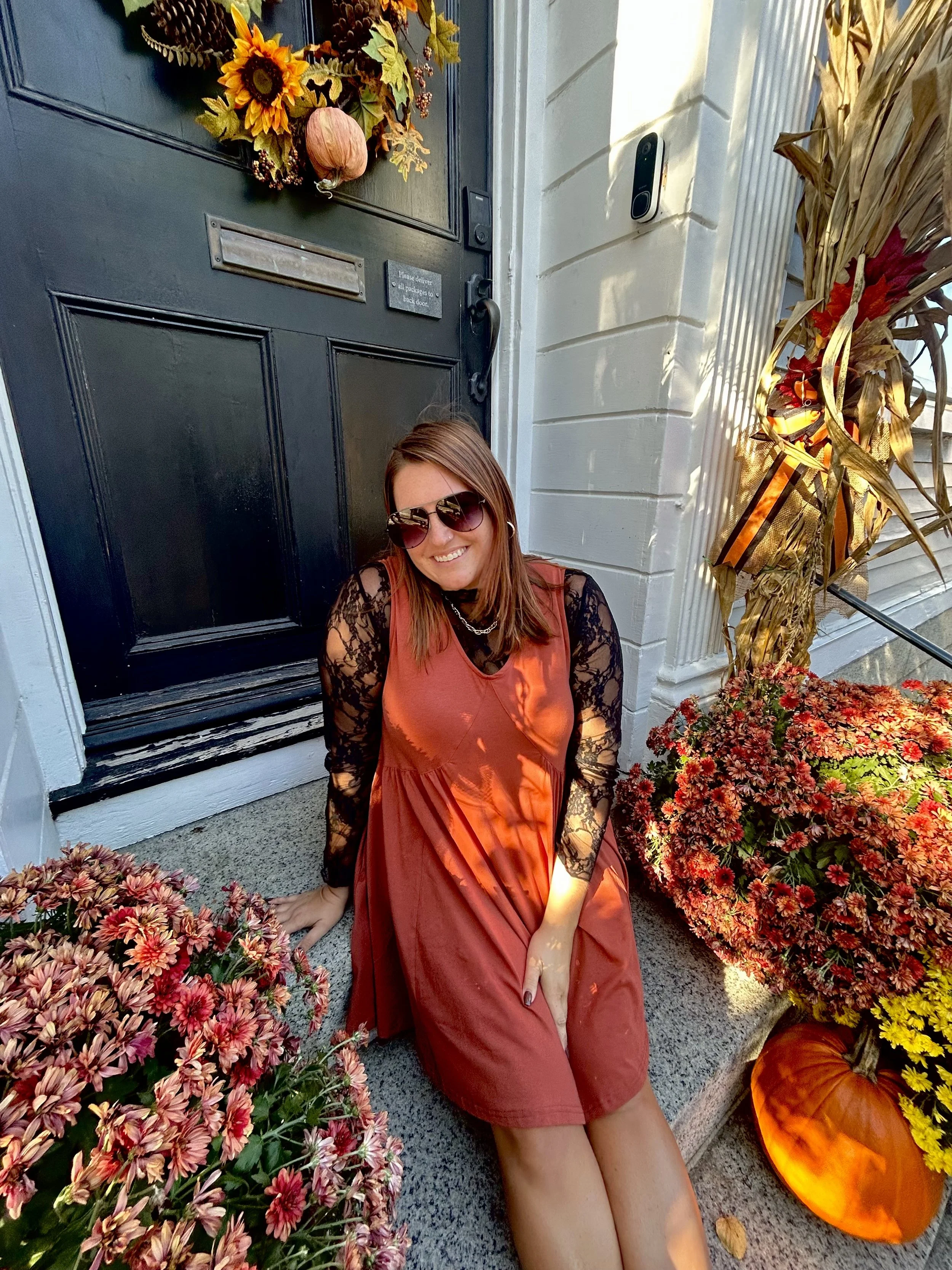 A woman with brown hair wearing sunglasses and an orange dress with black lace sleeves, sitting on the ground outside a house. She is smiling at the camera, surrounded by fall-themed decorations including pumpkins, yellow and orange plants, and a black front door decorated with a wreath of sunflowers, leaves, berries, and a small pumpkin. The house has white siding and a black mailbox, and the scene is illuminated by sunlight.