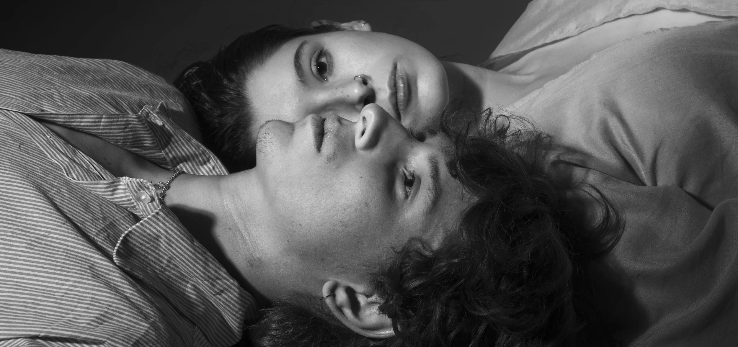 Two young women lying down close together, one resting her head on the other's shoulder, both looking at the camera in black and white.