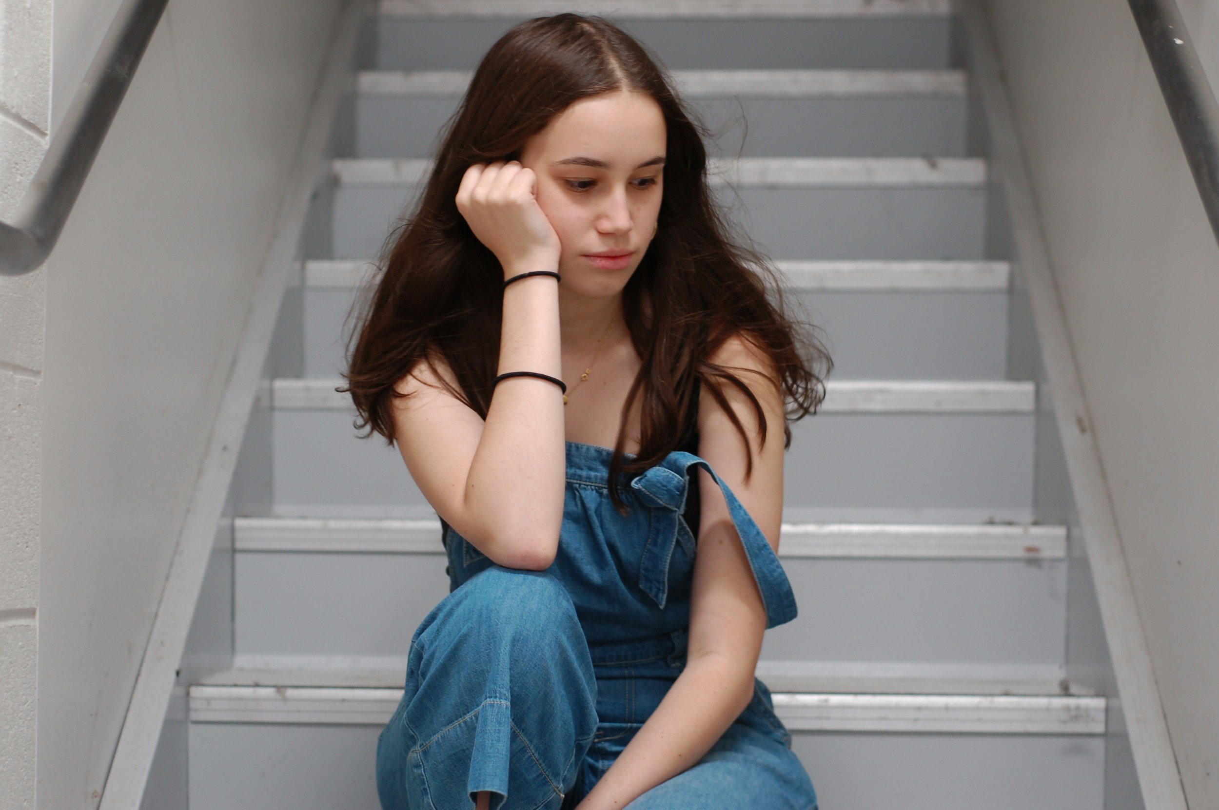 A young woman with long brown hair sitting on a staircase with her head resting on her hand, looking downward, appearing contemplative or sad.