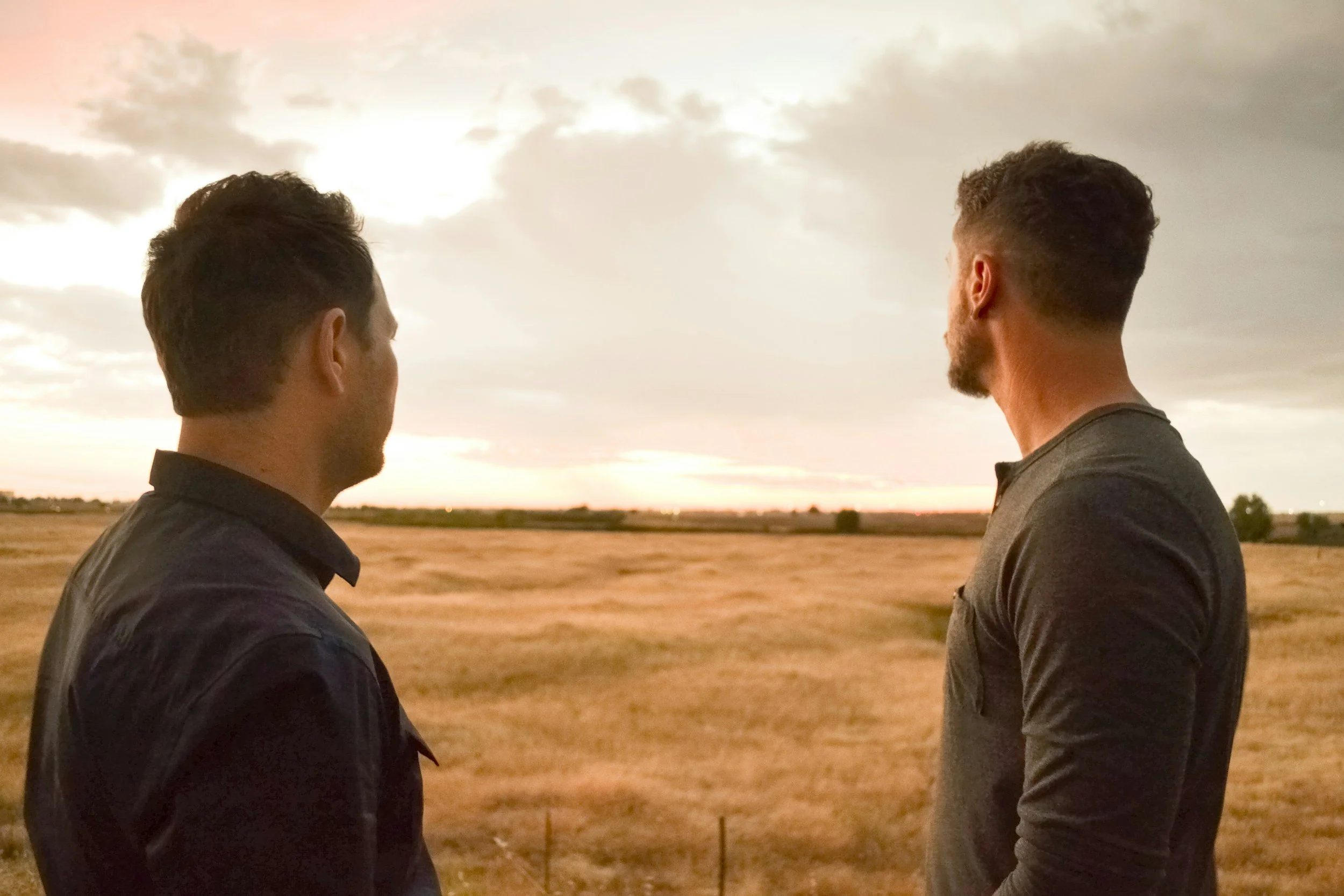 Two men standing outdoors at sunset, facing each other, with a field and cloudy sky in the background.