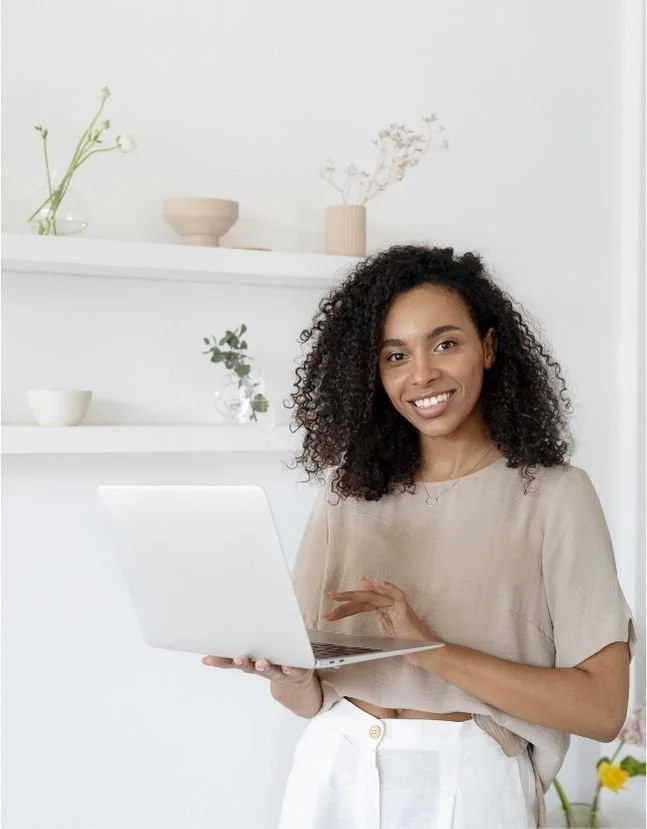 Smiling woman holding a silver laptop in a bright minimalistic room with white shelves, vases, and plants