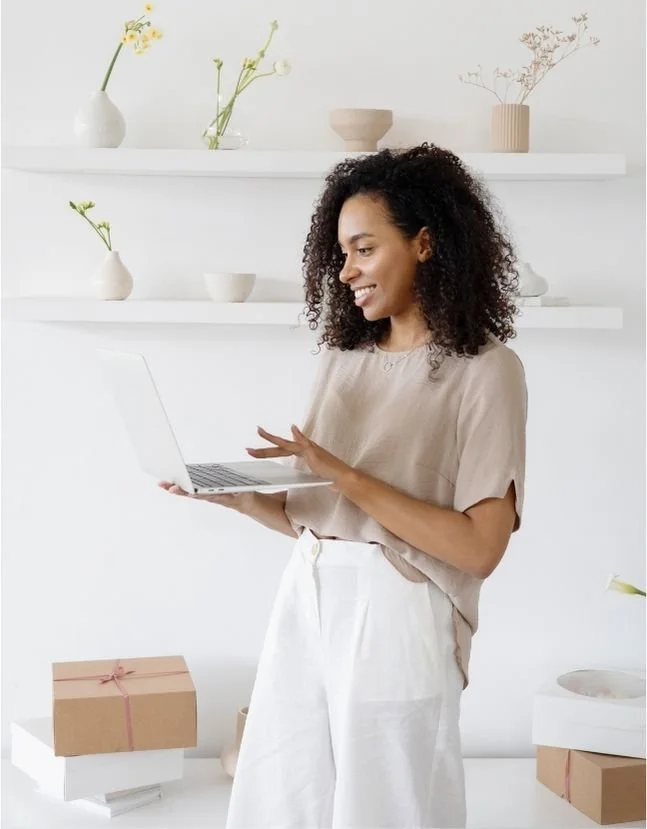 A woman with curly hair smiling and using a laptop in a minimalist room with white walls, shelves with vases and plants, and presents or boxes on a table.