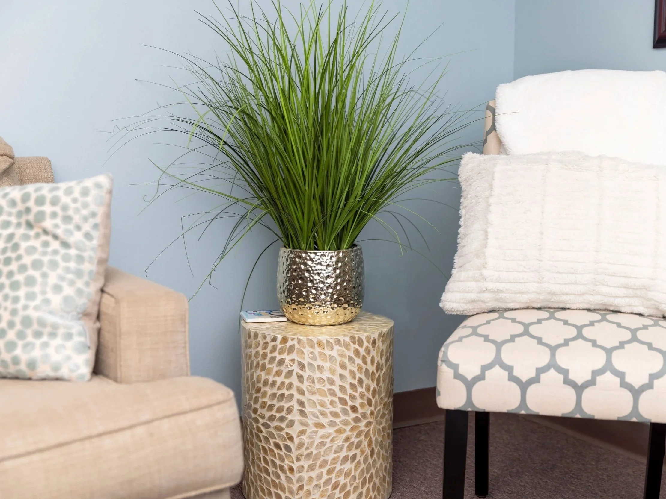 A tall decorative vase with a hammered metallic top and a textured beige bottom holding a large green plant with long slender leaves, placed on a round side table in a living room with beige and white furniture and light blue walls.
