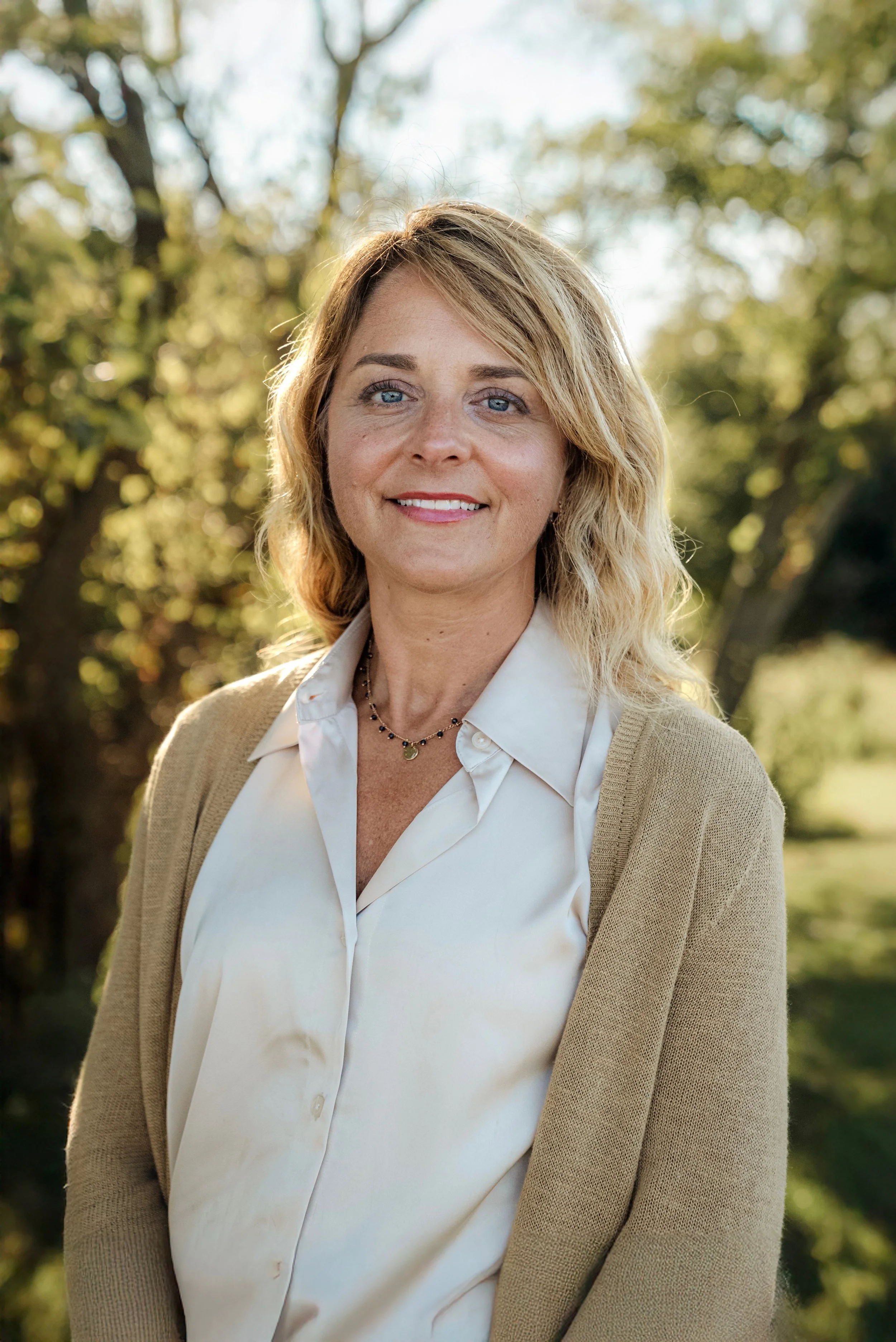 A woman with blonde hair, blue eyes, and light makeup smiling outdoors in front of trees with green and yellow leaves, dressed in a white blouse and beige cardigan, with a necklace.